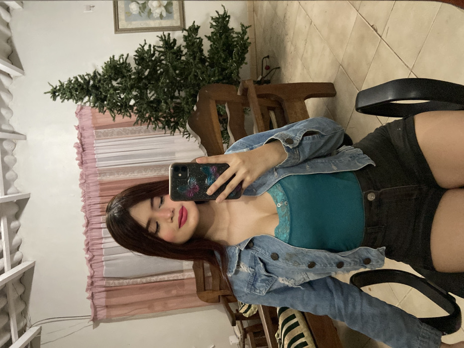 A young woman with long, dark hair takes a selfie. She wears a teal, lace-trimmed top, a distressed denim jacket, and black shorts. The phone she uses has a dark case with colorful designs. In the background, there's a wall with light pink curtains, a decorative shelf, and a Christmas tree-like plant. A wooden chair and a black office chair are also visible. The floor is tiled, and a framed picture hangs on the wall.
