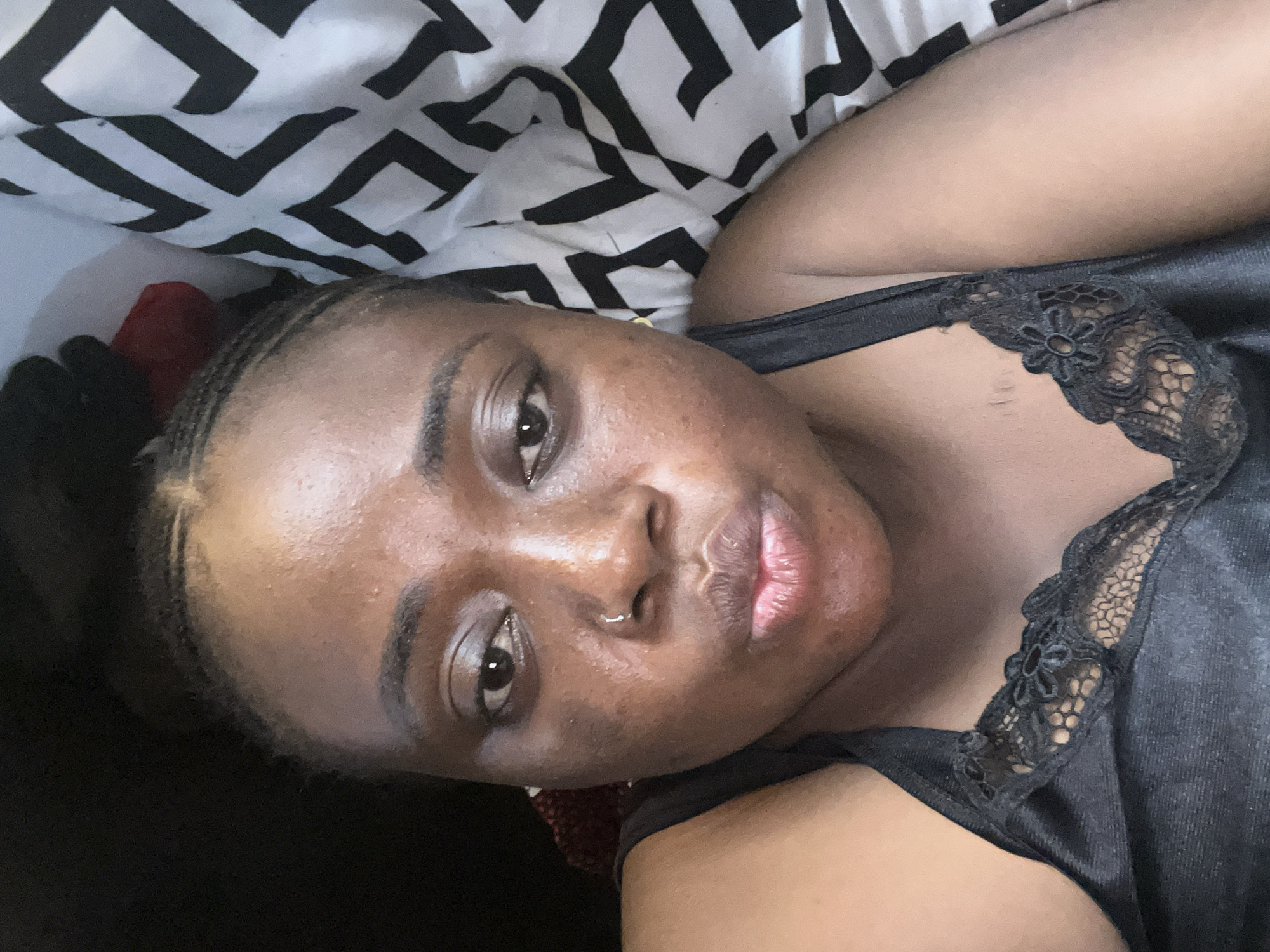A close-up, angled selfie of a woman with dark skin. She has her hair braided back from her face. She has a nose ring in her left nostril. She is wearing a black satin top with lace trim around the neckline. The background shows a patterned black and white pillowcase. No location in Abuja, Nigeria, is discernible from the visual information provided.