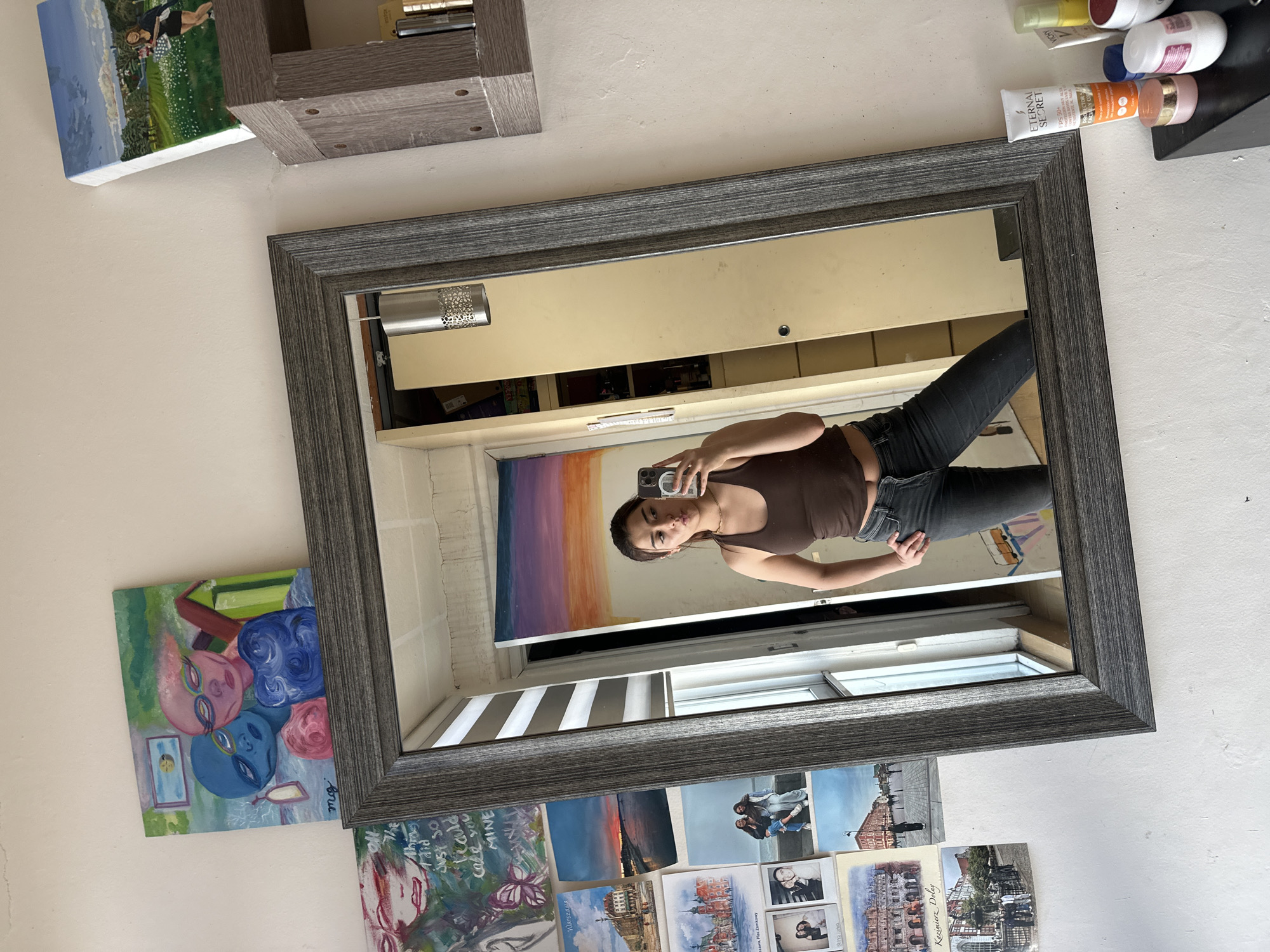 This image captures an indoor mirror selfie, rotated 90 degrees counter-clockwise. The central subject is a young woman, reflected in a large rectangular mirror with a dark, textured frame. She has dark hair, wears a brown tank top and dark jeans, and is holding a smartphone to take the picture, displaying a neutral expression with a slight pout.
The light-colored wall surrounding the mirror is adorned with various decorations. To the upper left, a grey wooden floating shelf holds a painting of a person in a green landscape. Below this shelf, a square painting features stylized blue and pink alien-like faces and is signed "mg." Along the bottom edge of the frame, numerous postcards and small prints are taped to the wall, showcasing landscapes, cityscapes (including one labeled "Warszawa" and another "Kazimierz Dolny"), and personal photographs. A small artwork among them contains fragmented text, including "I did JUST SO Beauld cake you MINE."
Inside the mirror's reflection, behind the woman, a light-colored cabinet or wardrobe is visible, along with a window fitted with horizontal blinds. A modern light fixture with a decorative, perforated metal shade is mounted above the cabinet. To the upper right, outside the mirror's frame, a black wall-mounted shelf holds several cosmetic products, including a tube marked "ETERNAL SECRET FPS 50+" and a bottle of "VICHY." The scene appears to be illuminated by natural daylight entering through the window. The setting is contextualized as Puebla, Mexico.
