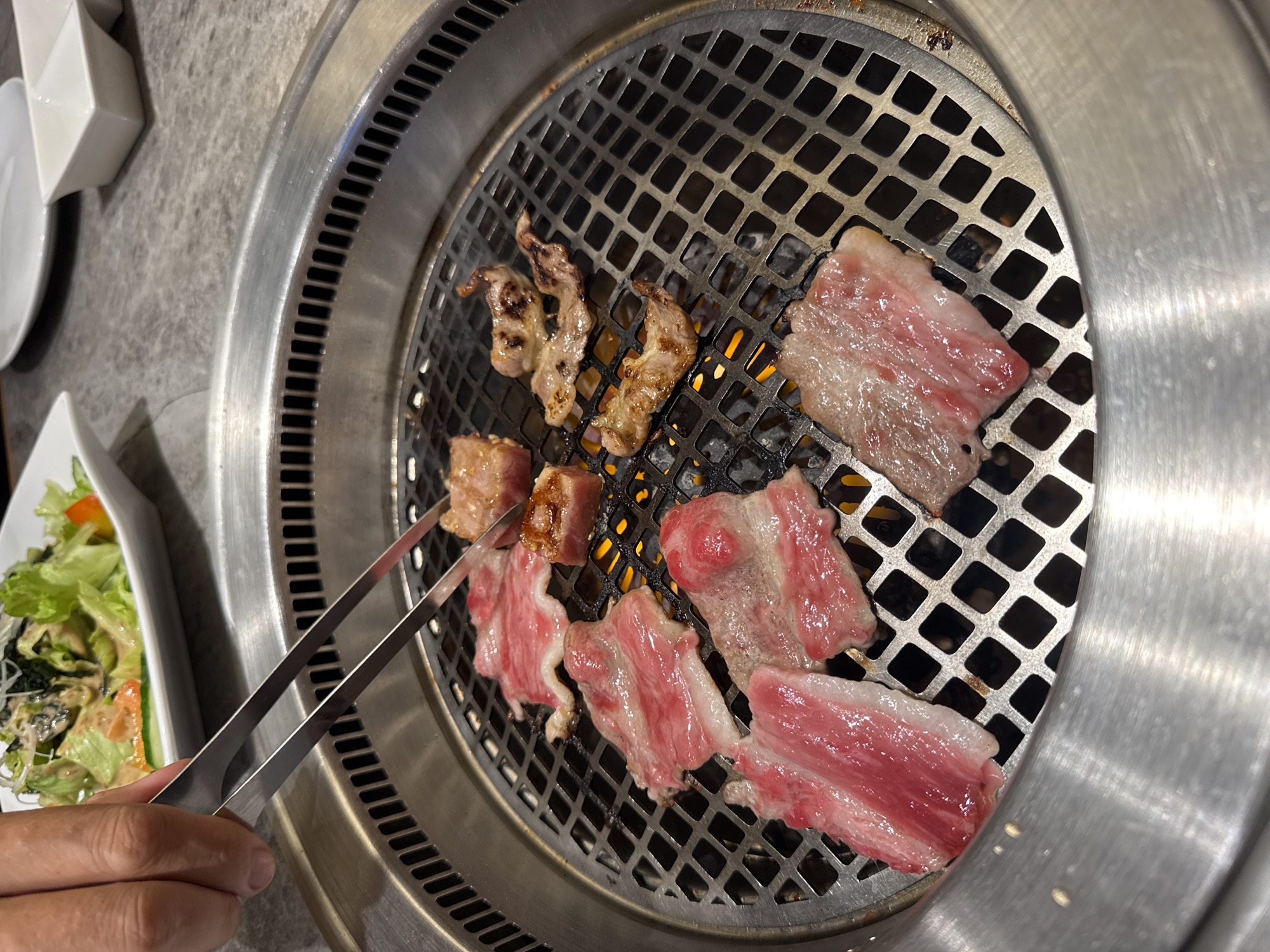 The image presents a close-up, overhead view of a tabletop BBQ grill in an indoor restaurant setting, located in Kuta Selatan, Indonesia. The main activity is grilling various cuts of meat.
The central subject is a circular, stainless steel grill with a metal grate, underneath which glowing heat from charcoal or gas flames is visible. Several pieces of meat, predominantly thinly sliced and appearing to be beef with visible marbling (streaks of fat), are cooking on the grate. Some pieces are still raw and pink, while others are partially cooked, showing browning and slight charring. A few pieces are more cooked and appear curled from the heat.
In the lower-left corner, a person's hand is visible, holding a pair of metal tongs, actively turning or placing meat on the grill. To the left of the grill, a white rectangular plate holds a side salad composed of green lettuce, shredded carrots, and what appears to be dark seaweed, all topped with a light dressing. Other white ceramic dishes are partially visible in the background, resting on a textured, gray table surface.
The scene captures an interactive dining experience, typical of yakiniku or Korean BBQ restaurants, where patrons cook their own food at the table. No specific time of day is discernible, and there is no visible text in the image.