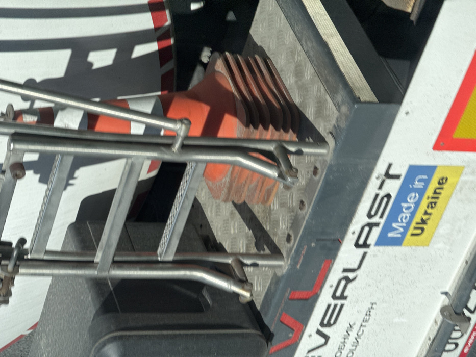 The image displays a close-up, elevated view of the rear or side platform of a utility vehicle, situated on Peremohy Street, Ukraine. The platform itself is constructed from metallic diamond plate. A polished metal ladder with multiple rungs is securely mounted on this platform. To the right of the ladder, a stack of at least six orange traffic cones, each featuring white reflective bands, is visible, with the uppermost cone fully exposed. A dark, rectangular object, possibly a storage box or toolbox, is partially visible on the platform's left side. Directly below the platform, on the vehicle's white body, the brand name "EVERLAST" is printed in black lettering, with the "V" in red, accompanied by additional, partially visible Cyrillic text. Below the "EVERLAST" branding, a blue and yellow sticker, resembling the Ukrainian flag, prominently displays the text "Made in Ukraine." A partial red and yellow reflective marker is visible at the bottom right of the frame. Strong, defined shadows are cast across the white vehicle surface and the platform, indicating bright, direct sunlight. No individuals are present in the scene.