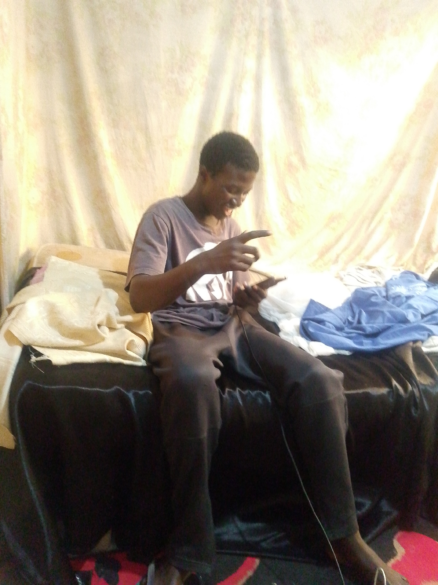 A young Black male, appearing to be in his late teens or early twenties, is sitting on a bed. He is wearing a dark purple t-shirt with white lettering and dark gray sweatpants. He is holding a mobile phone in his right hand and appears to be interacting with it, looking down at the screen with a slight smile. His left hand is raised with his index finger extended, possibly pointing at the phone or gesturing. The bed is covered with dark fabric and has several items on it, including a folded light-colored blanket or towel on the left and a crumpled blue cloth on the right. Behind the bed is a draped fabric, likely curtains, with a pale yellow hue and visible folds. The lighting suggests an indoor environment, possibly with natural light coming from the right, creating some overexposure in that area. The image does not contain specific landmarks or details that would allow for precise identification of the location within Jos, Nigeria.