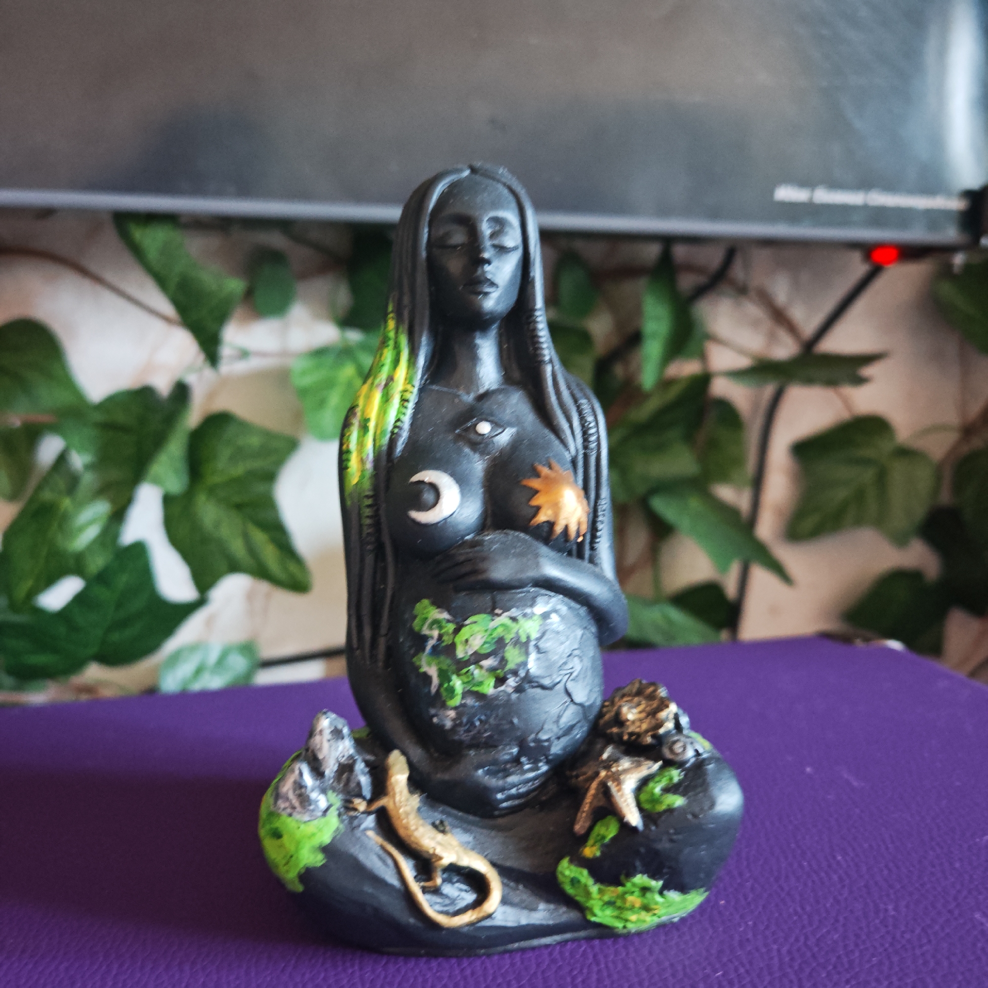 A black, pregnant goddess statue is centered on a purple surface. The statue is adorned with painted details, including a crescent moon and sun on the breasts, a third eye on the forehead, and green foliage on the stomach and base. A gold lizard and starfish decorate the base. Behind the statue is a TV and a wall covered with green vines. This image does not contain information to support a location in Rostov, Russia.
