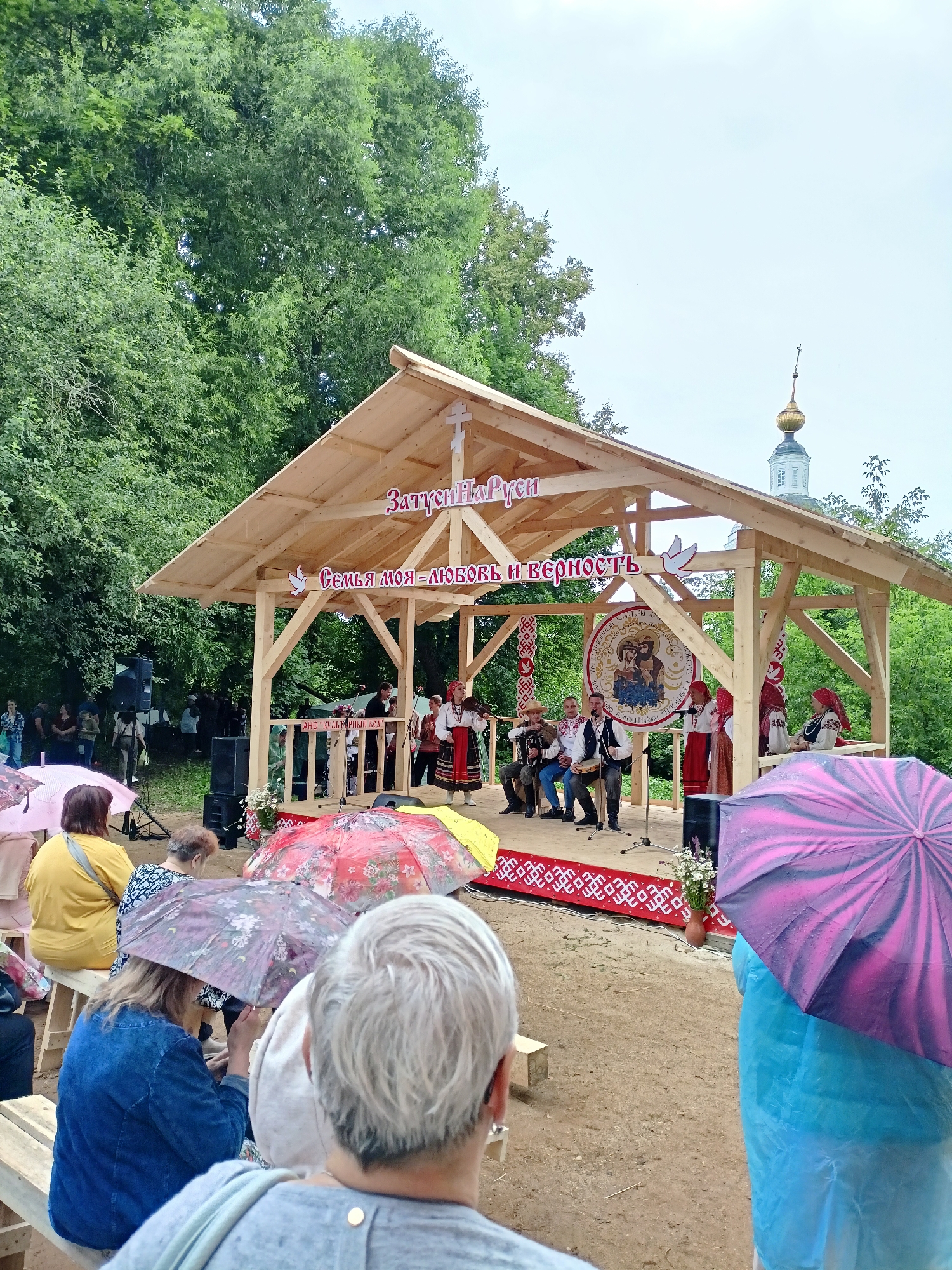 An outdoor event is taking place under a wooden canopy. A group of people in traditional clothing are performing on a stage. The canopy has a wooden roof supported by posts, and a banner with Russian text reads "Семья моя – любовь и верность" (My family is love and faithfulness). There is a circular emblem on the stage with an image of what appears to be a religious icon. The ground in front of the stage is dirt. Several people are seated in the audience, some holding umbrellas due to light rain. In the background, lush green trees frame the scene, and a glimpse of a church steeple can be seen. The overall atmosphere suggests a cultural or religious festival.