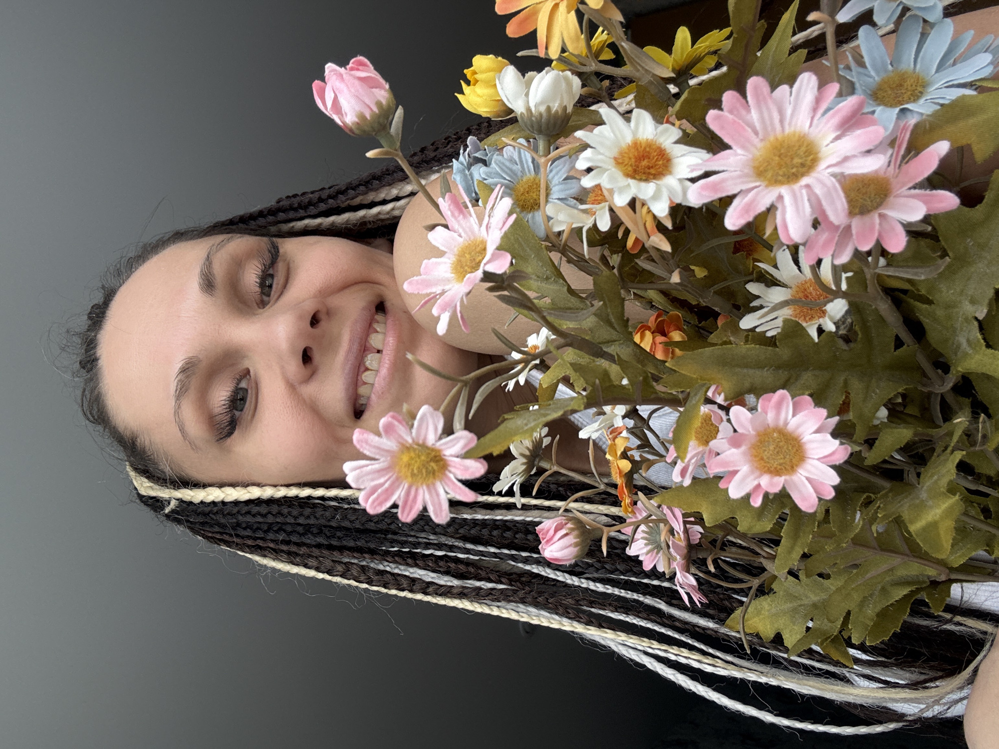 A woman with braided hair, colored brown, white, and black, is shown holding a large bouquet of artificial flowers in the foreground. The flowers are a mix of colors including pink, white, yellow, and light blue daisies, intermingled with green leaves. The woman is smiling, and her face is partially obscured by the flowers. The background is a simple, neutral gray wall. The location is unknown, and there is no discernible link to Rusanovo, Russia.