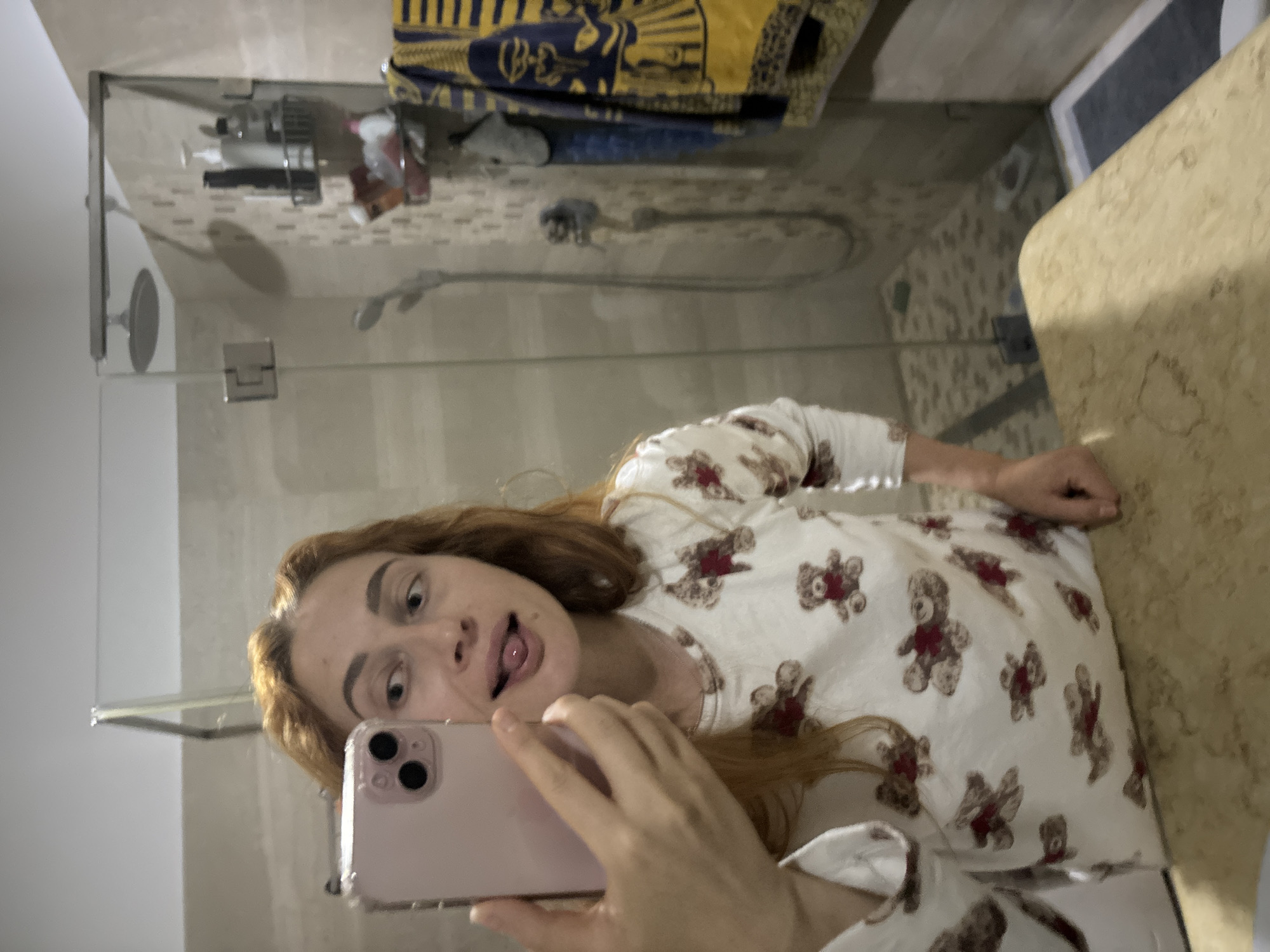 A person is taking a selfie in a bathroom. The person has red hair and is wearing a white pajama top with red teddy bear patterns. The person is sticking their tongue out. The bathroom has a glass shower door, a shower head, and a beige countertop. A yellow and blue towel hangs above the shower. The location is Budva, Montenegro.