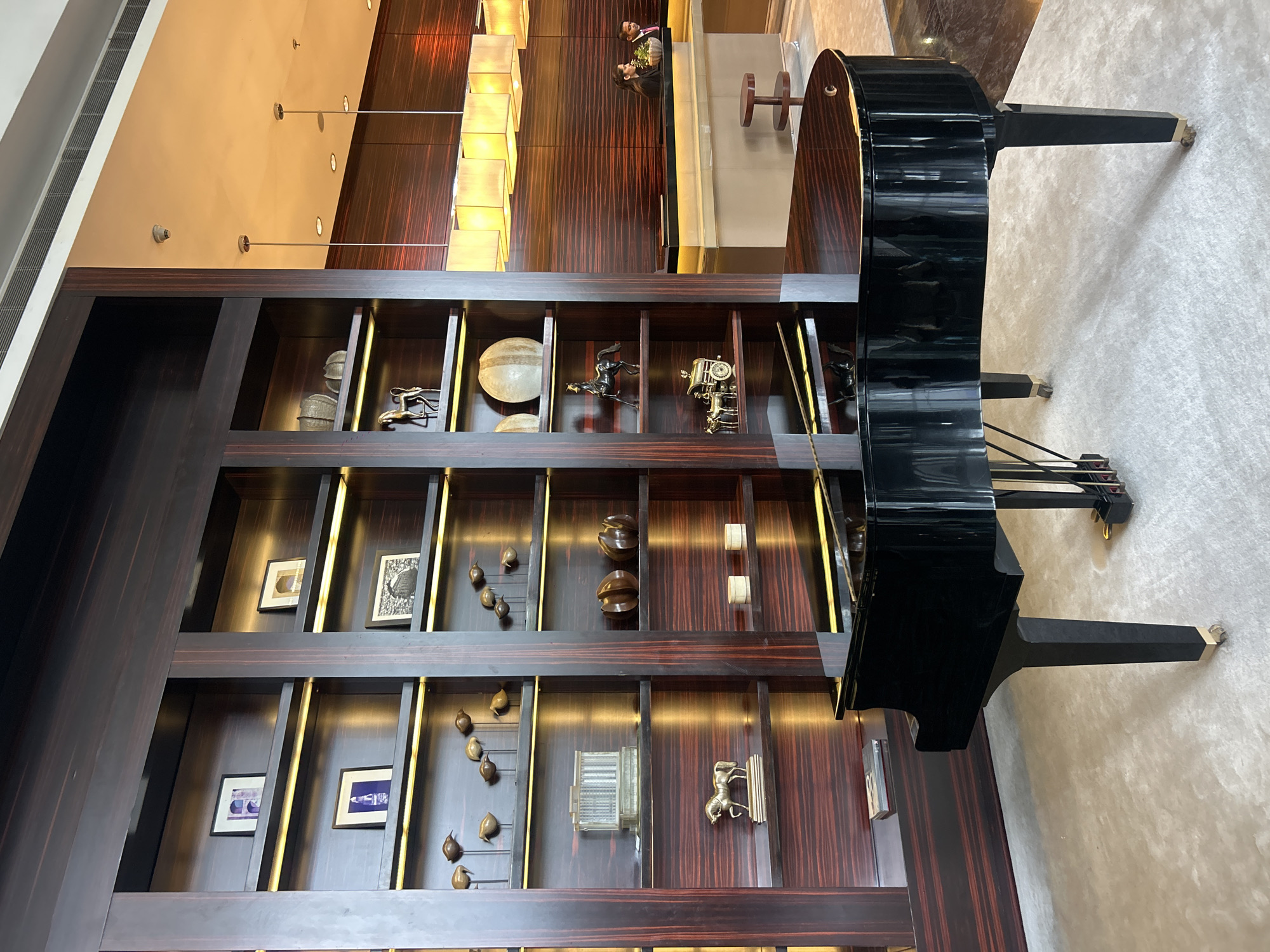 The image shows the lobby of the Conrad Pune, a luxury hotel located in Pune, India. In the foreground, a glossy black grand piano sits on a light-colored rug. Behind the piano is a tall, dark wood bookshelf filled with various decorative items, including framed pictures, sculptures of horses, and abstract pieces. The bookshelf is illuminated by warm, recessed lighting. To the right of the bookshelf, a reception desk is visible, with two hotel staff members, a woman and a man, attending to it. The reception desk is modern and sleek, with a dark countertop and light-colored cabinets. The walls behind the desk are paneled with dark wood, and a row of square pendant lights hangs from the ceiling, providing soft illumination. The overall ambiance is sophisticated and elegant, typical of a high-end hotel lobby.