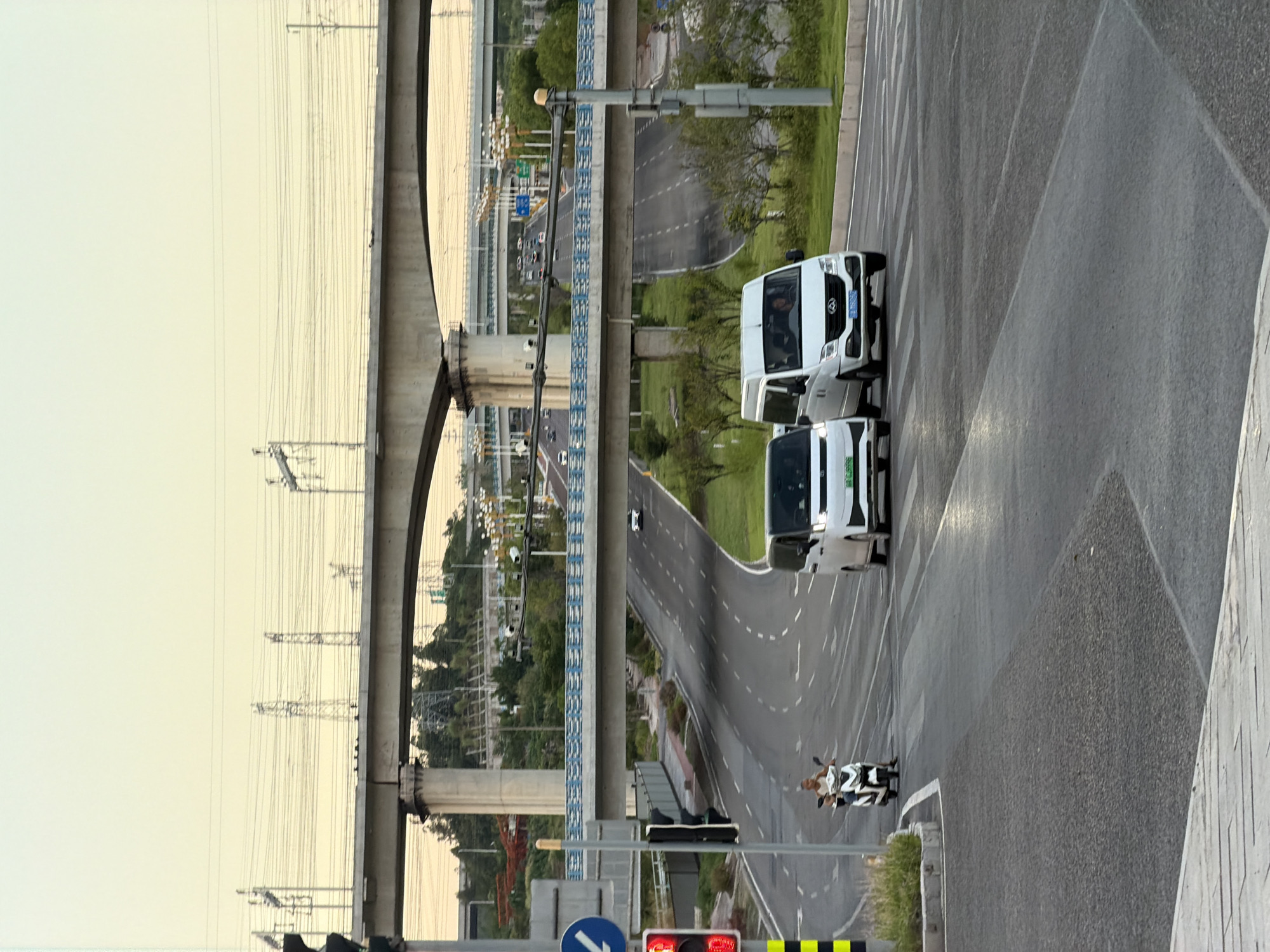 The image shows a road intersection in Jiangjin, China, with multiple elevated highways crossing overhead. In the foreground, a traffic light is visible on the left, displaying a red signal. A person is riding a white electric scooter across the intersection. Two white vans are also present, one in the left lane and one in the right lane. The environment is urban, with modern infrastructure, including overhead power lines and streetlights. The sky is overcast with a pale yellow hue, suggesting it may be early morning or late afternoon. The road surface is asphalt, and lane markings are clearly visible.