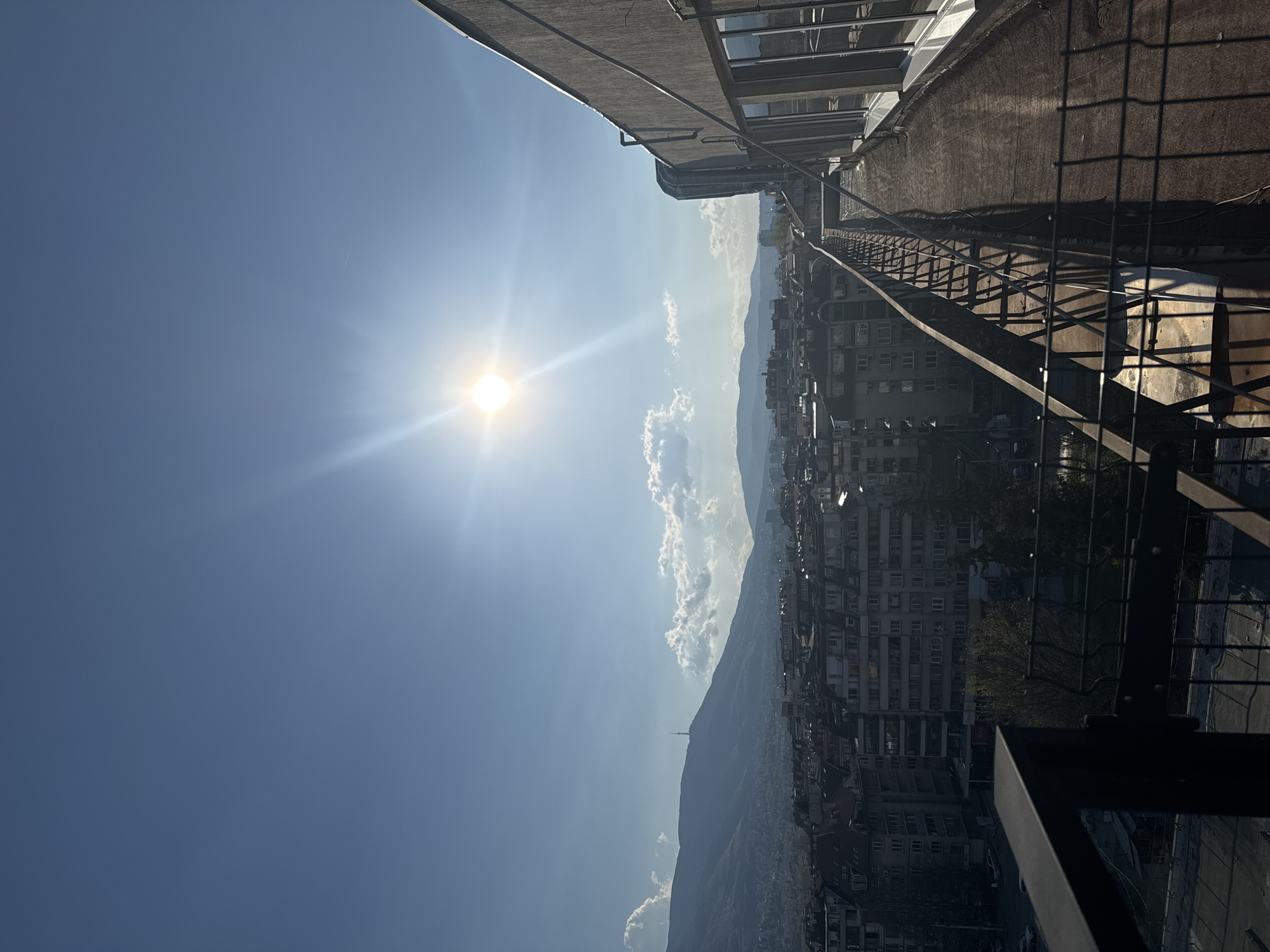 The image displays a panoramic view of a city nestled against a backdrop of mountains, under a bright, sunny sky. The photograph is taken from a high vantage point, likely a building's rooftop, looking out over the urban landscape.
In the foreground, on the right side of the horizontally oriented image, is a dark, industrial-style structure with a metal fence or railing, possibly part of the rooftop infrastructure from which the picture was captured. A flat, concrete surface, typical of a building roof, is also visible.
The midground reveals a dense urban sprawl, consisting of numerous multi-story buildings with visible windows and roofs, stretching across the frame. The architectural style suggests a city in Eastern Europe.
Dominating the background is a large mountain range, its peaks partially obscured by a light atmospheric haze. On one of the prominent mountain peaks, a distinct communication tower can be seen, which, given the provided context, is identifiable as the Kopitoto TV Tower on Vitosha mountain, confirming the location as Sofia, Bulgaria.
The sky is a vibrant, clear blue, adorned with several scattered white, fluffy clouds above the mountains. The sun is prominently featured in the upper left portion of the sky, emitting strong rays and lens flare, indicating a bright and clear day, likely midday or early afternoon. No individuals or discernible text are visible in the scene. The overall impression is one of an expansive urban setting meeting natural mountain beauty under brilliant sunshine.