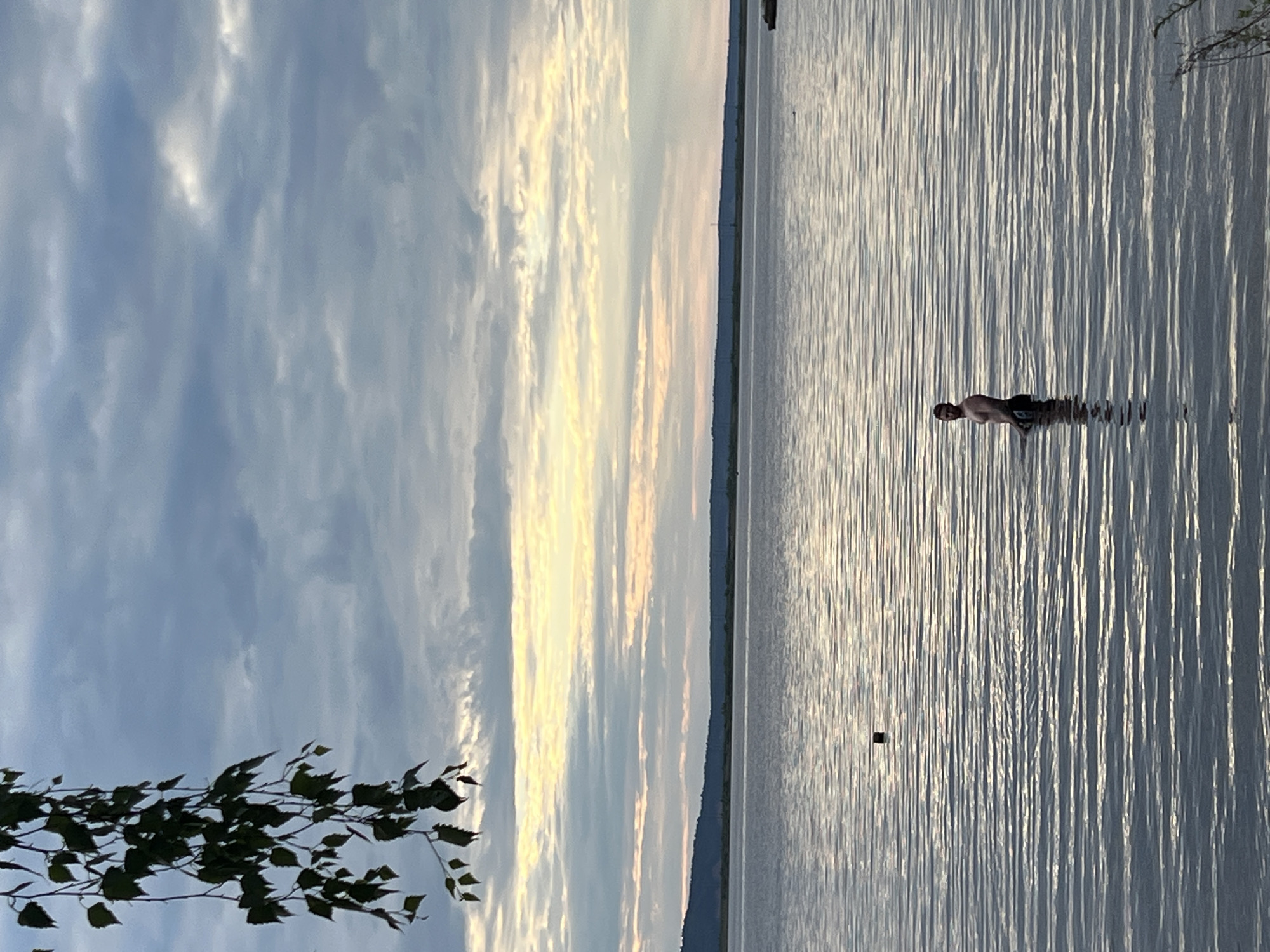 A man is standing in the calm, rippling water of a lake, with his torso and head visible above the surface. He is facing towards the left of the frame, with his arms down by his sides. The man is wearing dark-colored shorts. In the background, a distant, dark, tree-lined shoreline is visible under a cloudy sky with streaks of yellow and white light. A few dark green leaves hang from a branch in the upper left corner of the image. The overall scene suggests an outdoor environment, possibly during dusk or dawn, on the Kama River in Chaikovsky, Russia.