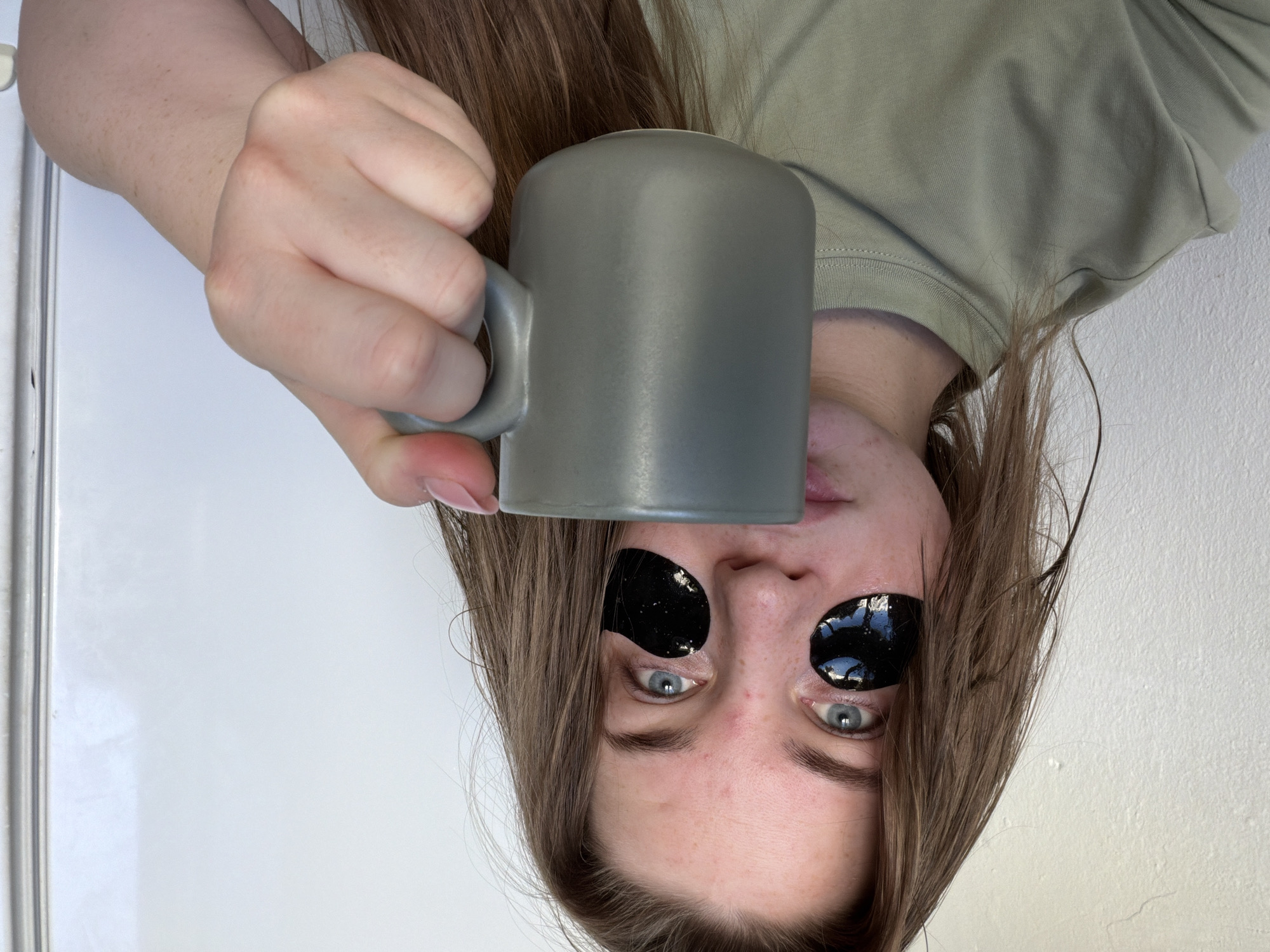 An upside-down close-up of a fair-skinned individual with long brown hair holding a gray mug. The individual is wearing black under-eye patches. The background is a plain white wall. The individual is wearing a green t-shirt. The location is possibly in the city of Chaikovsky, Russia.
