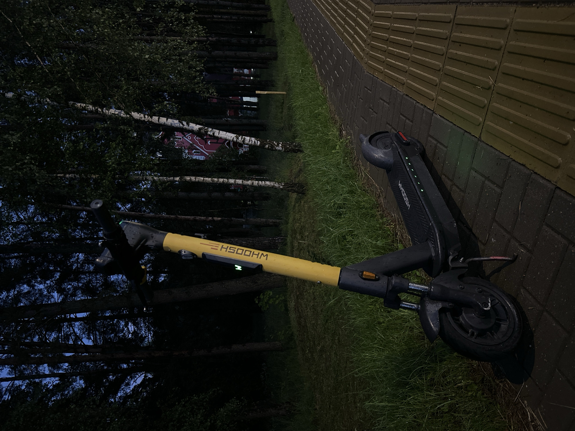 A yellow and black electric scooter is parked on a brick pathway next to a grassy area with trees. The scooter is the main subject, positioned in the center of the frame, angled slightly to the left. It is illuminated by ambient light, with its front wheel closest to the viewer. The pathway is paved with dark bricks, transitioning to textured yellow tiles on the right side of the frame. Behind the pathway and scooter, a dense line of trees forms a dark backdrop, with glimpses of a red building with arched windows visible through the foliage in the distance. The sky above is dark blue, indicating it is either dusk or night. The overall environment suggests an outdoor urban or suburban setting, possibly a park or a sidewalk in a residential area within Ждановіцкі сельскі Савет, Belarus. There are no people visible in the image.
