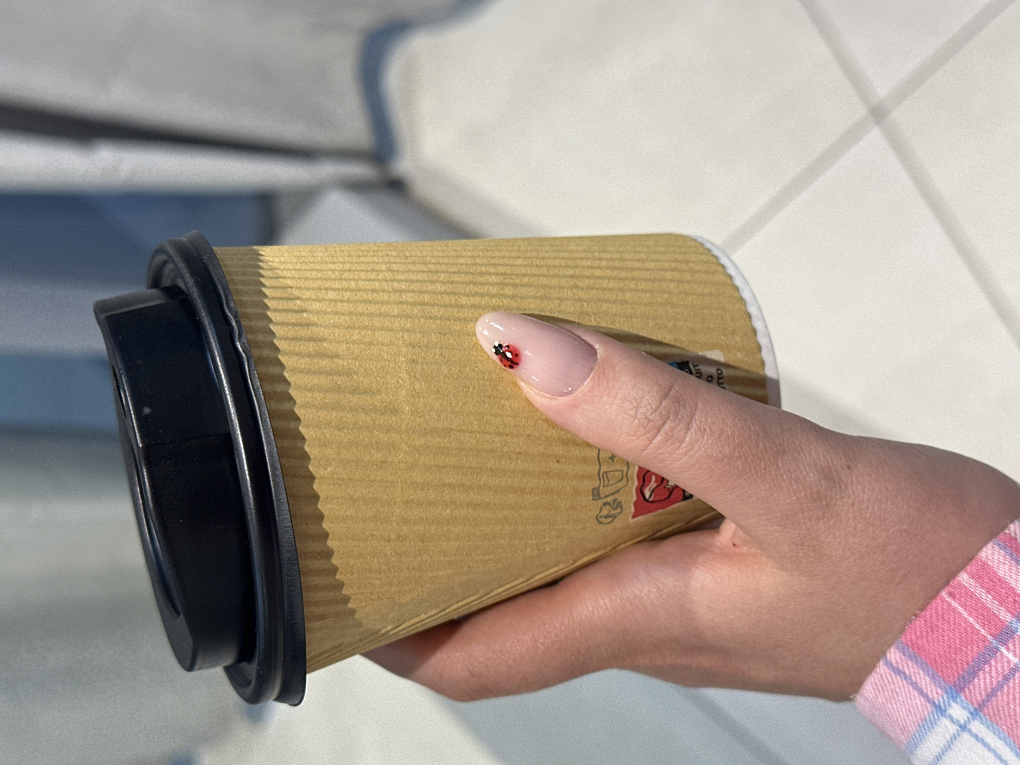 The image presents a close-up view of a person's hand holding a disposable coffee cup. The hand, likely female, has well-groomed nails, with a notable light pink or nude polish on the index finger adorned with a small, detailed red and black ladybug design. The person is wearing a plaid shirt, visible on the lower right, featuring a pattern of pink, white, and light blue squares.
The coffee cup is made of corrugated light brown cardboard, giving it a ribbed texture. It is fitted with a black plastic lid. Partial text and a red logo are visible on the cup's side; fragments like "UIT" and "TTO" are discernible, but the complete branding is obscured.
The background is softly out of focus, showing light-colored, possibly tiled or paneled surfaces, with subtle shadows that suggest bright, diffused lighting, indicative of daytime. The setting appears to be indoors or in a semi-enclosed environment, such as a cafe or office. The activity is simply holding a beverage. No specific emotions are expressed. The photo was taken in Šušanj, Montenegro.