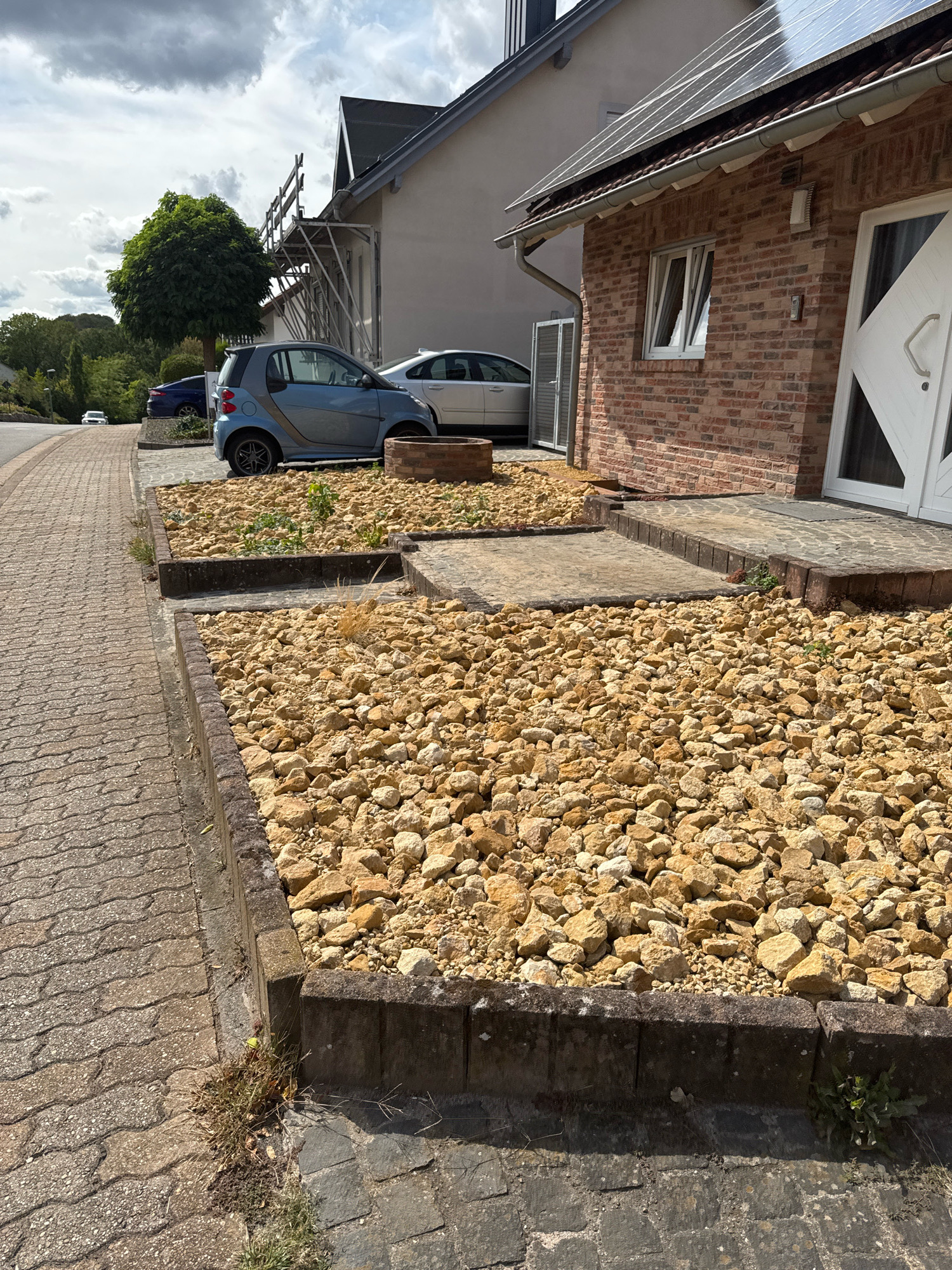 In Überherrn, Germany, on a cloudy day, a brick house with a dark tiled roof and solar panels is visible. A paved driveway leads to the entrance, which has a white door with diamond-shaped panes. Adjacent to the house is a garden bed filled with light brown gravel and scattered small green plants. To the left of the garden bed, a light blue Smart car is parked. Further down the street, a dark blue car and a white car are also parked. A paved sidewalk runs alongside the street, bordered by a low stone retaining wall in front of the garden bed. The environment appears to be a residential area with houses and trees.