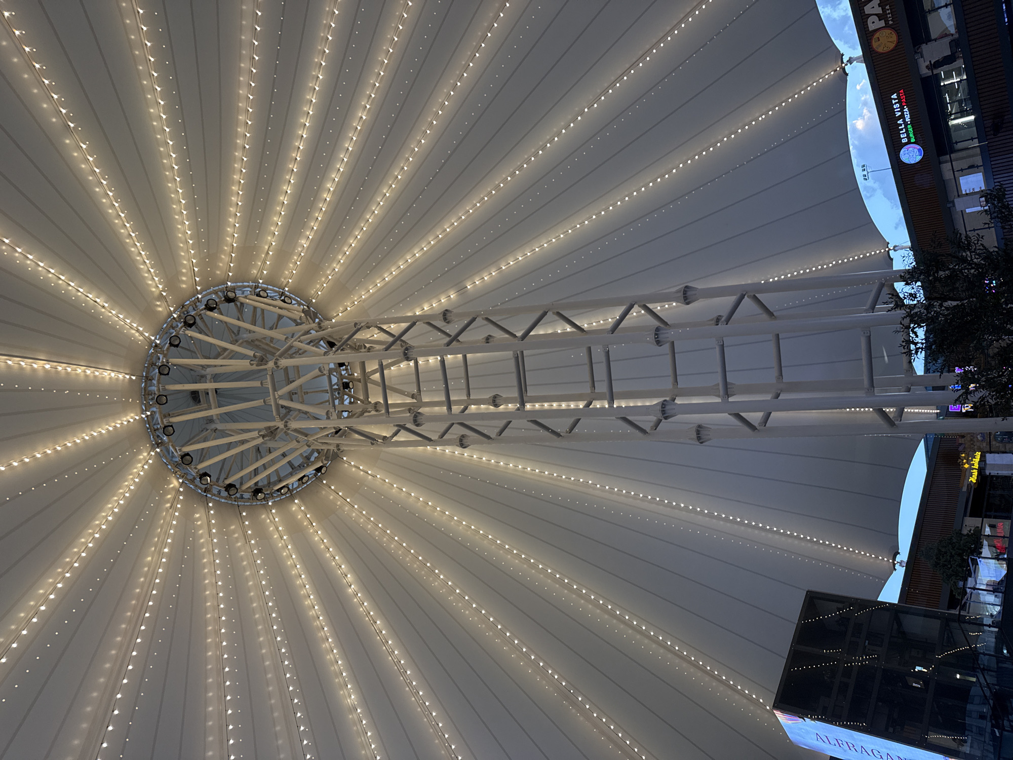 The image shows the underside of a large, white, tent-like structure with a central support tower. Numerous strings of small, warm-toned lights are attached to the fabric, creating a radial pattern emanating from the center. The fabric is stretched taut and features visible seams.
Below the tent structure, a multi-story building with various commercial storefronts is visible. One prominent sign reads "BELLA VISTA BURGER PIZZA PASTA". Another sign, partially visible, shows the word "PA" likely part of "PIZZA". A digital billboard on a separate structure displays the word "ALFAGAN". The overall setting appears to be an outdoor commercial or entertainment area.
Based on the visible signage and architectural style, this image is likely from the "Humoyun Plaza" shopping and entertainment complex located in Tashkent, Uzbekistan. The lighting suggests it is evening or night. No people are clearly discernible in the image.