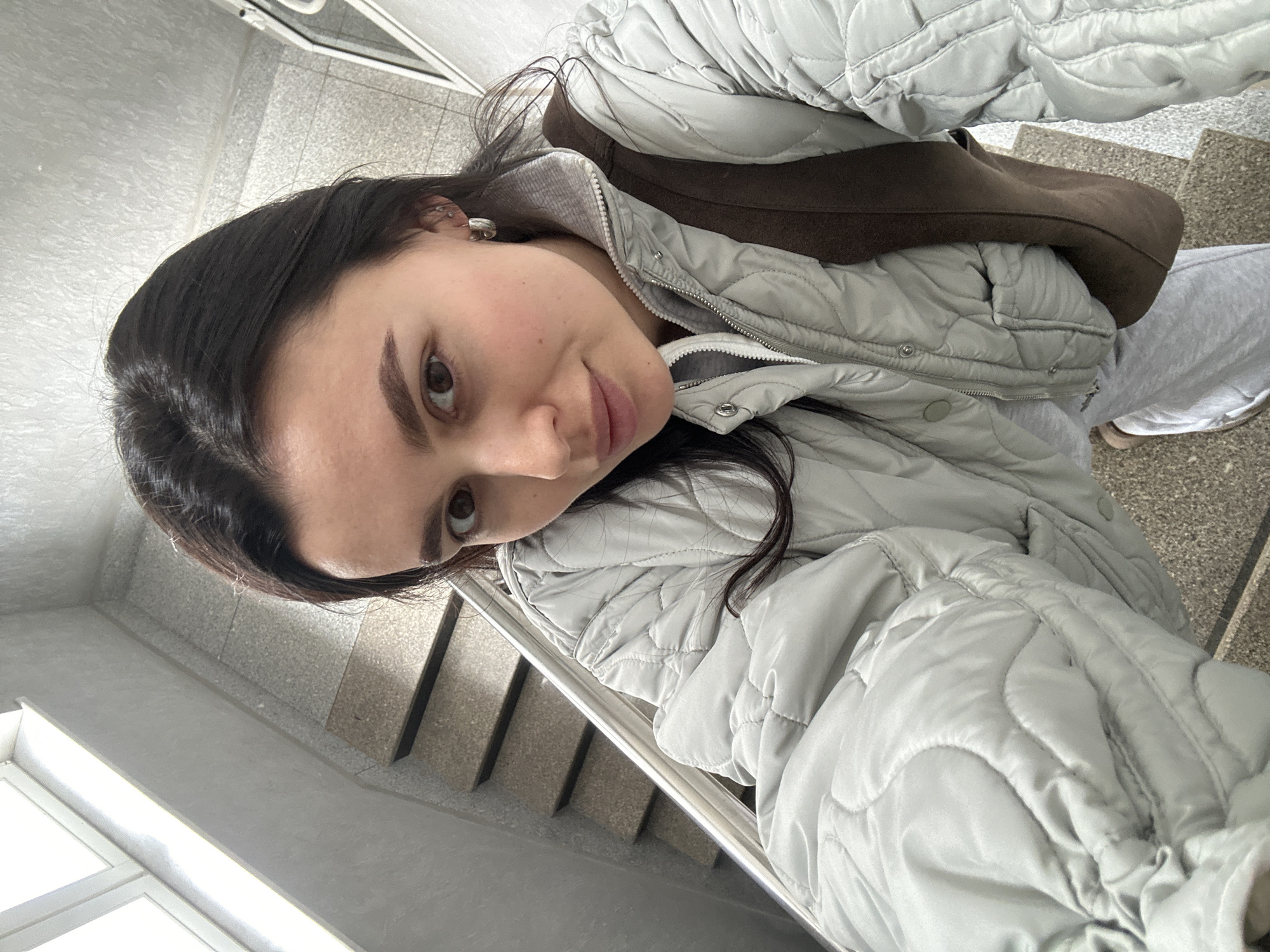 A young woman with dark hair is taking a selfie on a staircase. She is wearing a light grey puffer jacket and a brown bag is slung over her shoulder. The staircase appears to be indoors, with grey stone steps and a stainless-steel railing. The location is unspecified but could be the city of Chaikovsky, Russia.