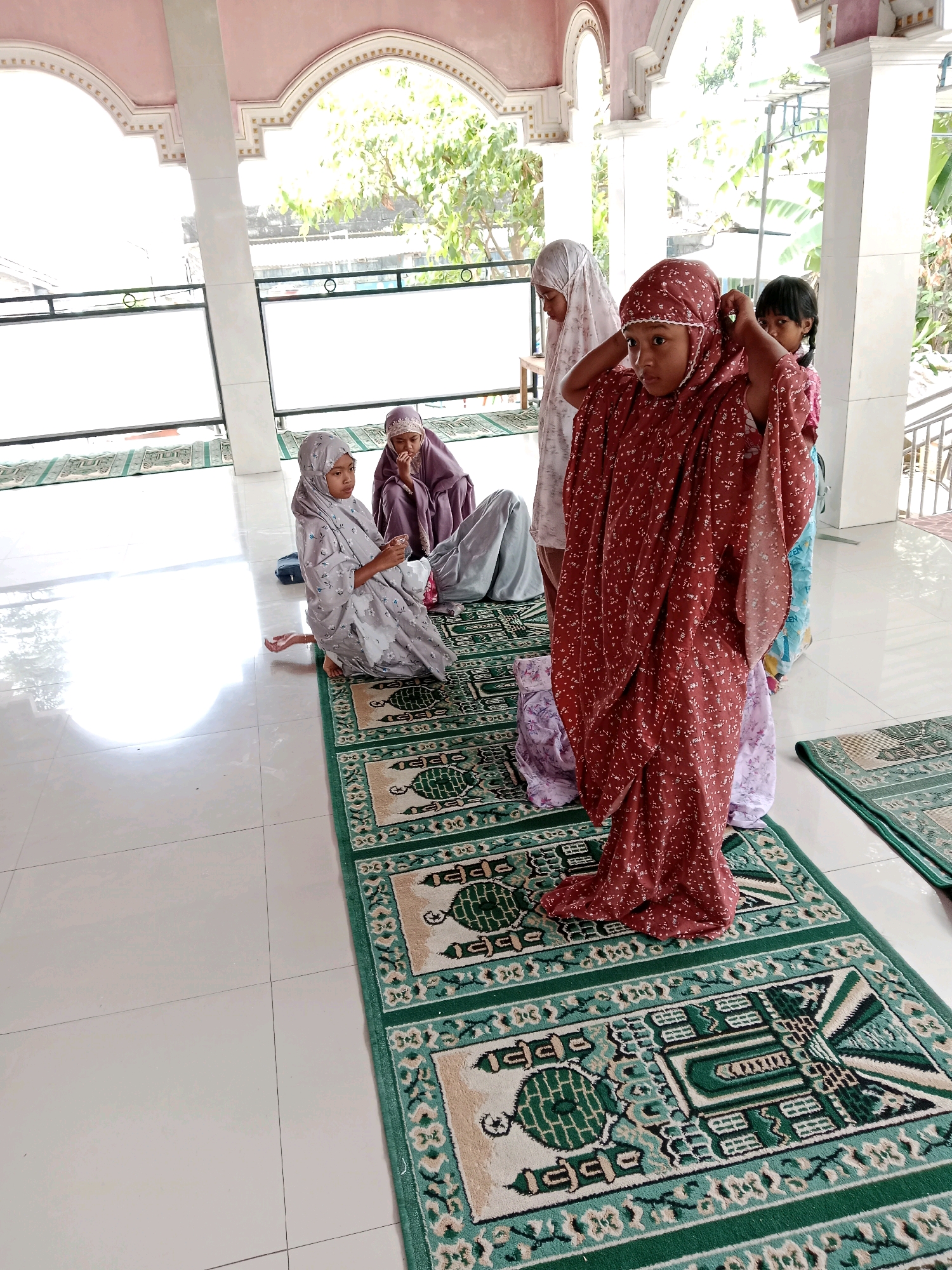 The image depicts a group of girls wearing hijabs and traditional Muslim attire, possibly preparing for prayer. They are situated indoors, on a tiled floor, with prayer rugs laid out. One girl in a red patterned garment is adjusting her hijab, while others are seated or standing nearby. The setting appears to be a communal space, possibly a mosque or a community hall, with architectural elements like arches and pillars visible. Sunlight streams in, illuminating the scene. The location is identified as Salatiga, Indonesia.