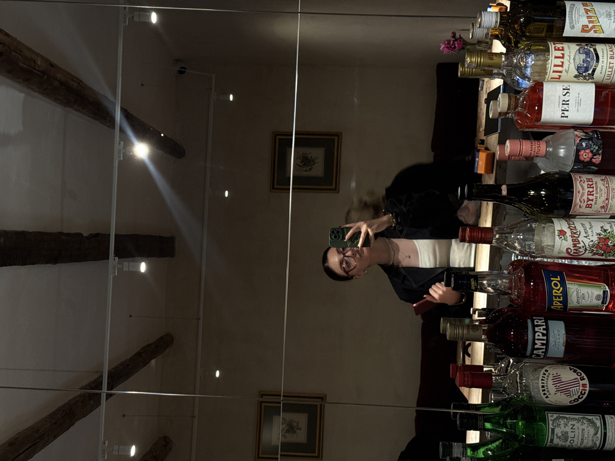 The image shows a mirror selfie taken in a room featuring a selection of alcoholic beverages. In the reflection, a woman with dark hair and glasses is holding a smartphone, presumably taking the photograph. She is wearing a black jacket and a light-colored top.
Behind the woman, the reflection reveals a room with beige walls and a framed picture hanging on the wall. The room also features a wooden beam across the ceiling.
On the right side of the image, a variety of bottles of alcohol are visible, including brands like Lillet, Per Se, Byrrh, Aperol, Campari, Cocchi Rosa, and Dolin, among others.
While the setting exhibits elements of a bar, the context is not explicitly confirmed, and location of the image is uncertain.