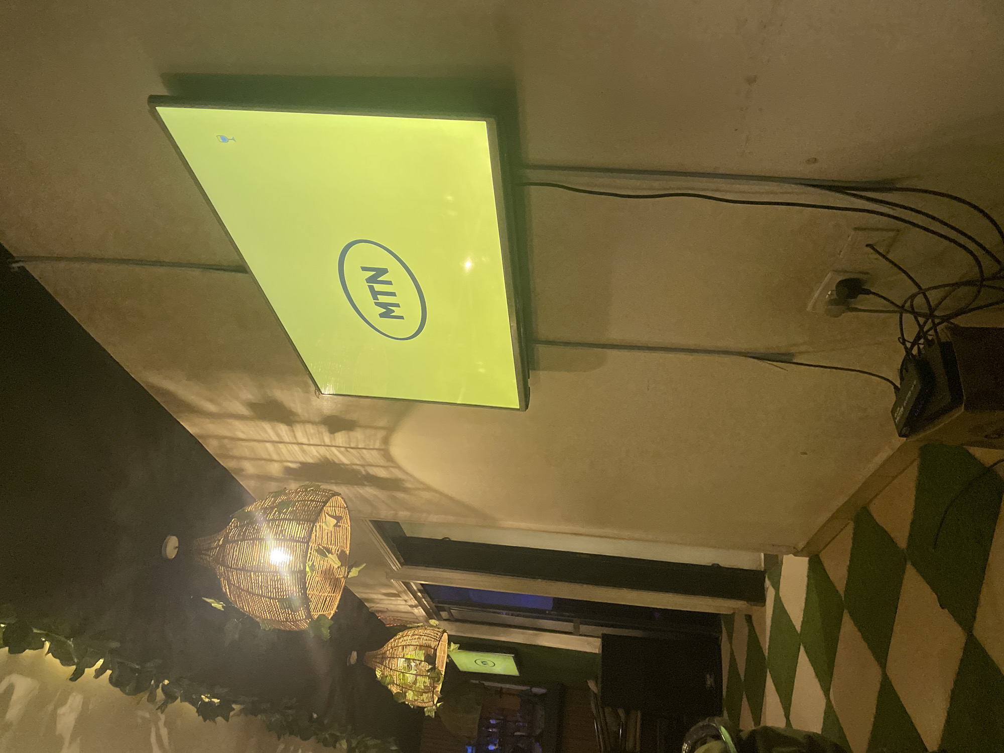 The image shows an interior setting, possibly a restaurant or bar in Jos, Nigeria, based on the presence of an MTN logo on a screen. The scene is illuminated with decorative woven pendant lights that have artificial vines attached. The walls are painted in a green and white diamond pattern. A window or doorway is visible in the background. There is an electrical outlet and cables running along the ceiling.