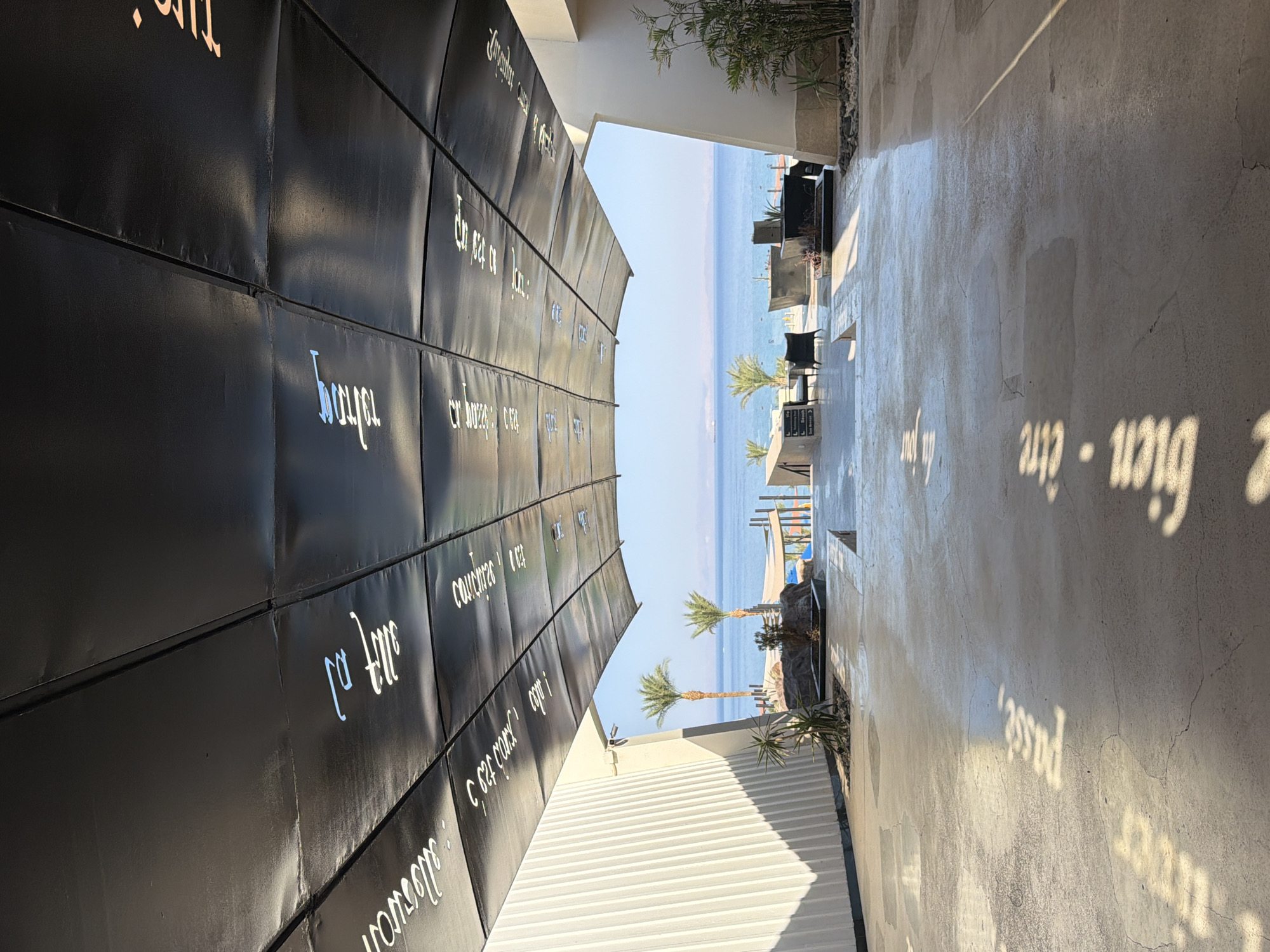 The image depicts a covered walkway leading to an outdoor area with a view of the Red Sea in Dahab, Egypt. The walkway's ceiling is made of dark panels with white text, possibly quotes or lyrics, written in a stylized script. The floor of the walkway is concrete and reflects sunlight, creating dappled patterns.
In the distance, beyond the walkway, the Red Sea stretches out under a clear blue sky. The water appears calm, with a few boats visible on the horizon. On the shore, there are palm trees and outdoor seating areas with modern furniture. Some of these areas appear to be near a boardwalk or pier.
To the left of the walkway, a textured wall with vertical lines is visible, casting a strong shadow on the concrete floor. To the right, there are some plants and rocks, along with architectural elements of a building. The overall scene suggests a resort or a public space designed for relaxation and enjoying the coastal scenery. There are no people visible in the image.
