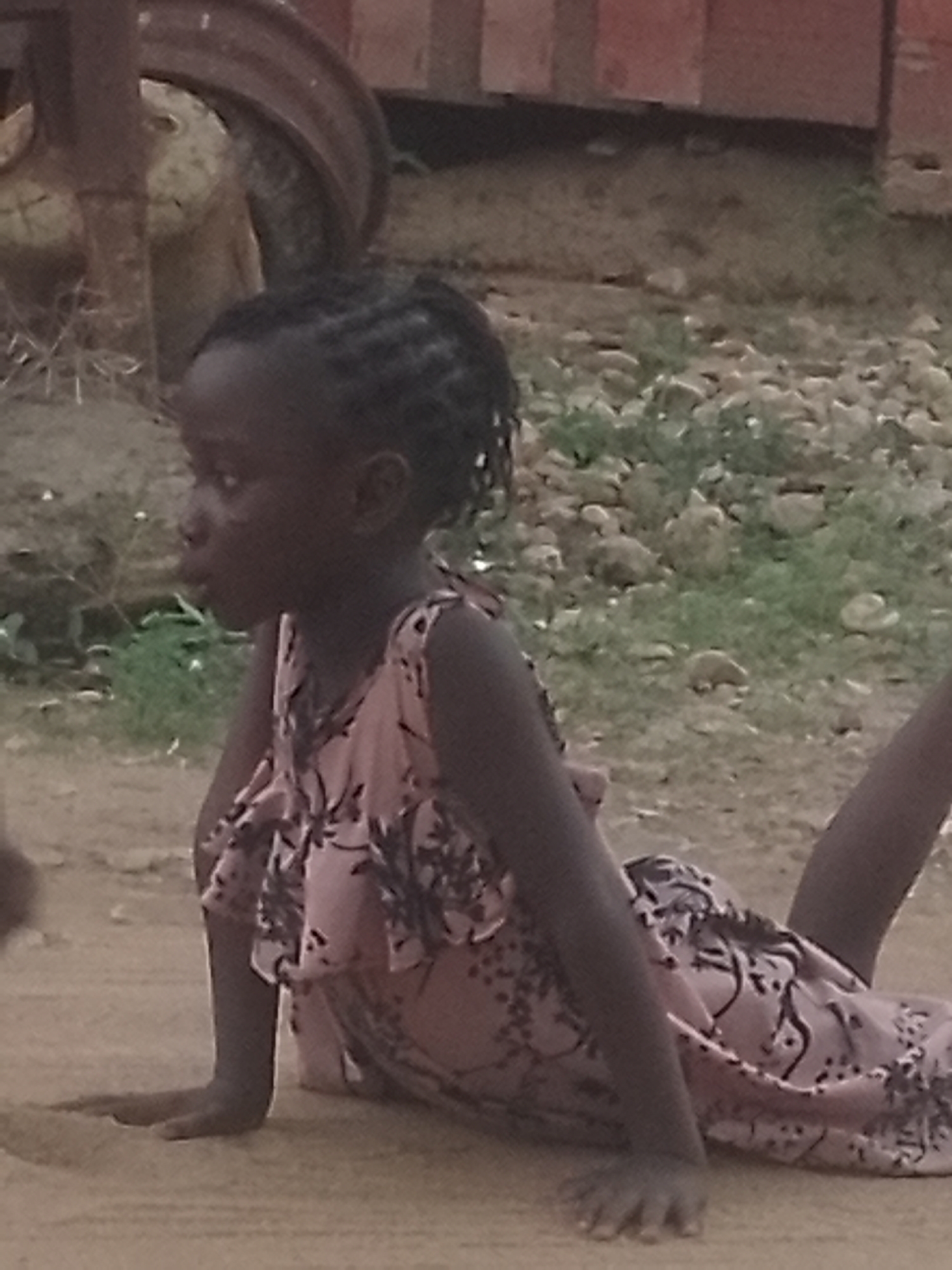 A young, dark-skinned girl with braided hair lies prone on a dirt surface. She supports herself on her forearms, lifting her torso and head. She wears a light pink dress with dark floral patterns. Behind her, the background consists of a mix of dirt, grass, and small stones, possibly a yard or outdoor area. A partially visible rusty wheel or tire is situated to the upper left. The location is likely Adankolo, Nigeria, as specified.