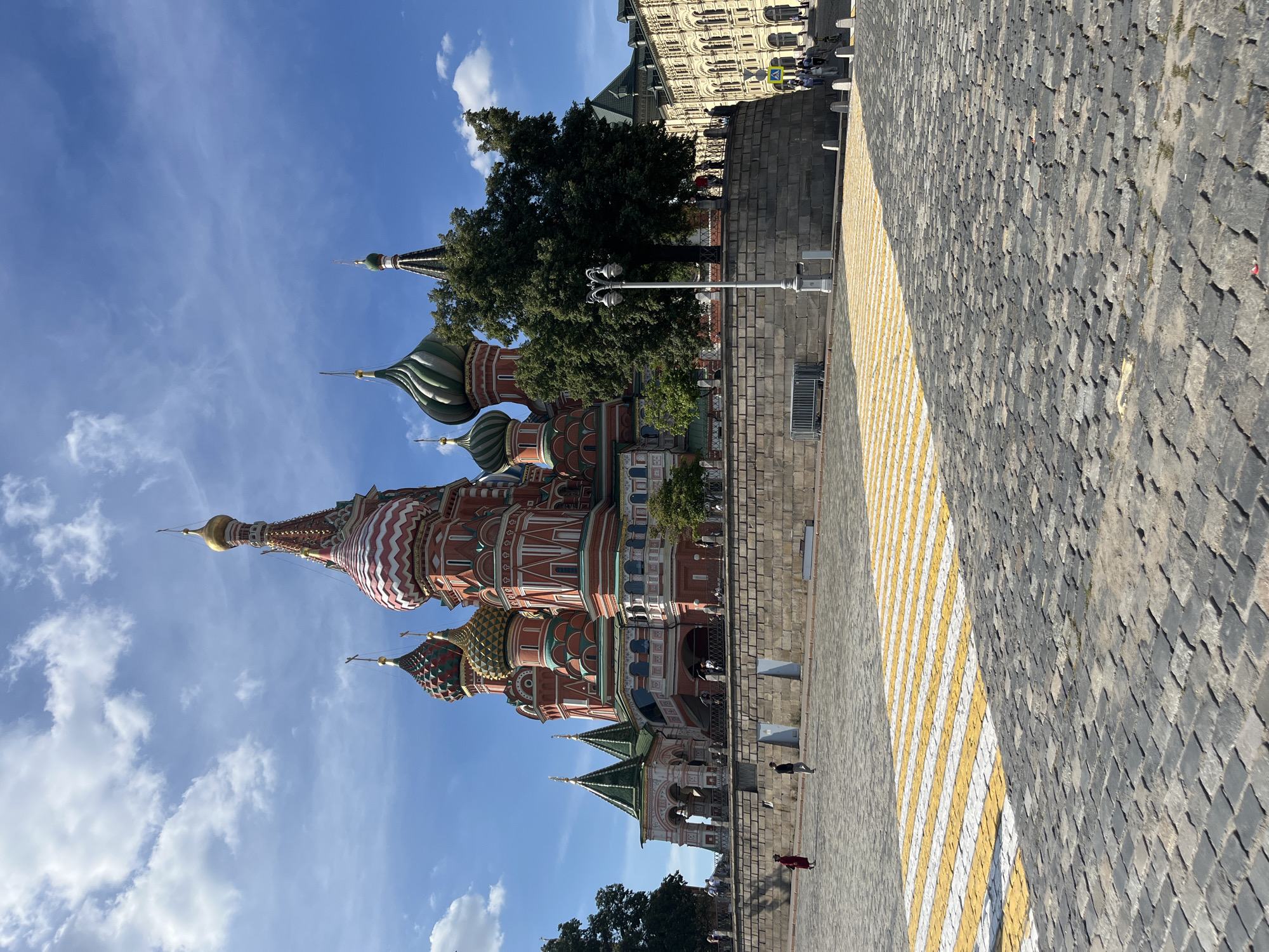 This image captures Saint Basil's Cathedral, a famous landmark located in Red Square, Moscow, Russia. The cathedral is characterized by its vibrant colors, onion domes, and intricate architectural details. The foreground features a cobblestone plaza with a yellow and white striped crosswalk. Several people are visible walking in the plaza and on the street in the background. Trees and other buildings, including a large, tan structure with arched windows, are also part of the scene. The sky is blue with scattered white clouds. The lighting suggests it is daytime.