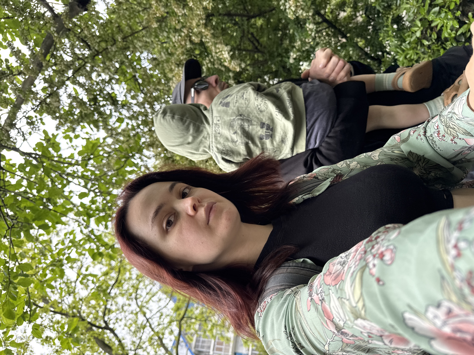 A ground-level, tilted shot shows a woman with reddish-brown hair in the foreground, looking directly at the camera. She wears a black top and a floral print green blouse. Behind her lies a man wearing sunglasses and a baseball cap, with a green hoodie over a black shirt. A child is lying next to him, with small feet and tan shoes visible. The background is filled with a dense canopy of trees. Based on the architectural details and the distinct flora, the location is most likely a park in the city of Sochi, Russia. The image captures a scene of relaxation or repose.