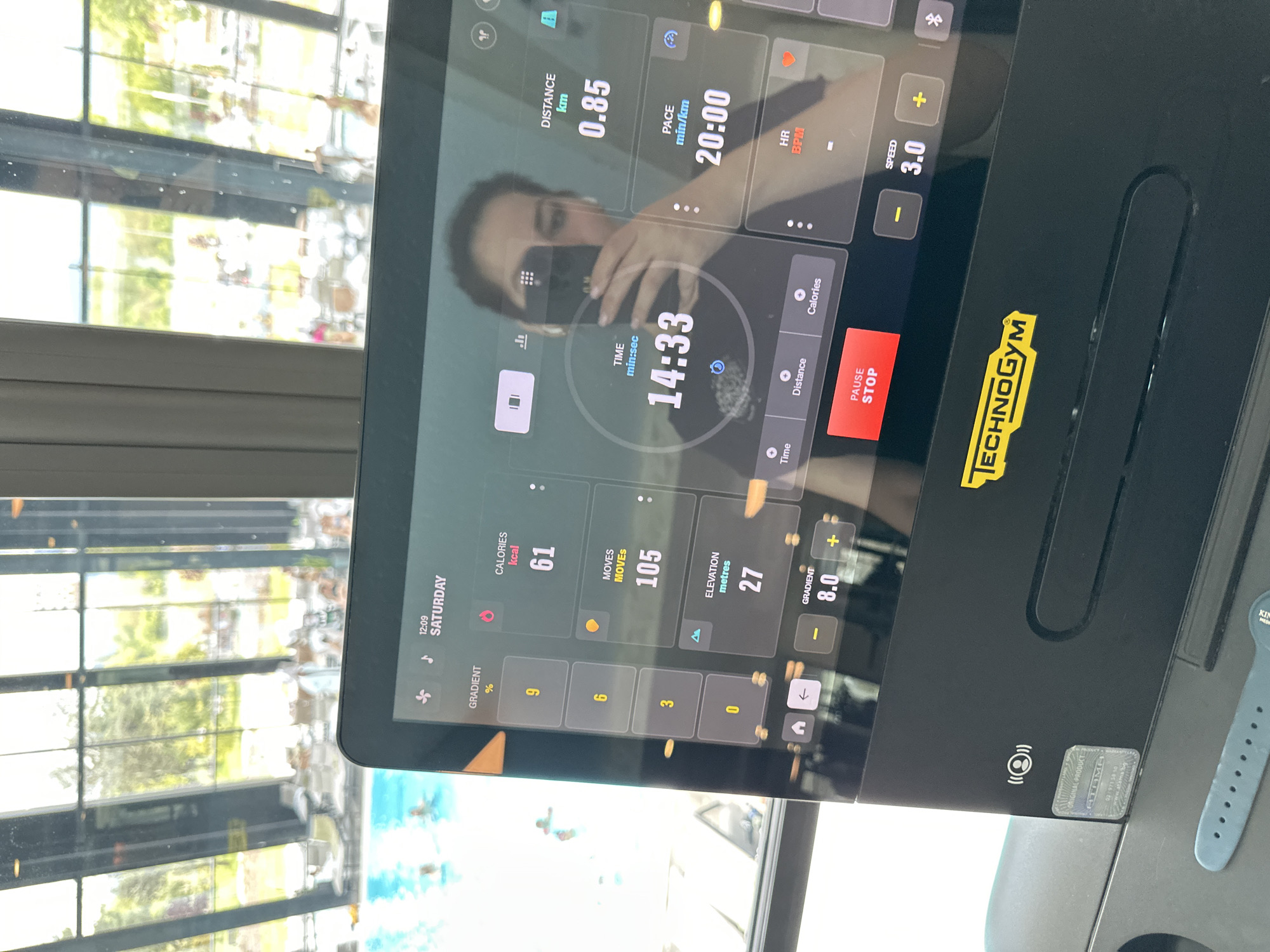 The image shows a close-up of a Technogym treadmill display screen with a reflection of a person holding a mobile phone, likely taking a picture. The screen displays workout metrics: a distance of 0.85 km, a pace of 20:00 minutes per kilometer, a time of 14 minutes and 33 seconds, and 61 calories burned. The treadmill speed is set to 3.0, and the gradient (incline) is at 8.0, though the display shows buttons for incline percentages of 9, 6, 3, and 0. The date shown is Saturday at 12:09. The reflection in the screen reveals the person is indoors, with a window behind them showing an exterior view of trees and buildings. The environment appears to be a gym or a location with exercise equipment and a view of the outdoors. It is not possible to determine the location in the city of Kazanlak, Bulgaria, from the provided image.
