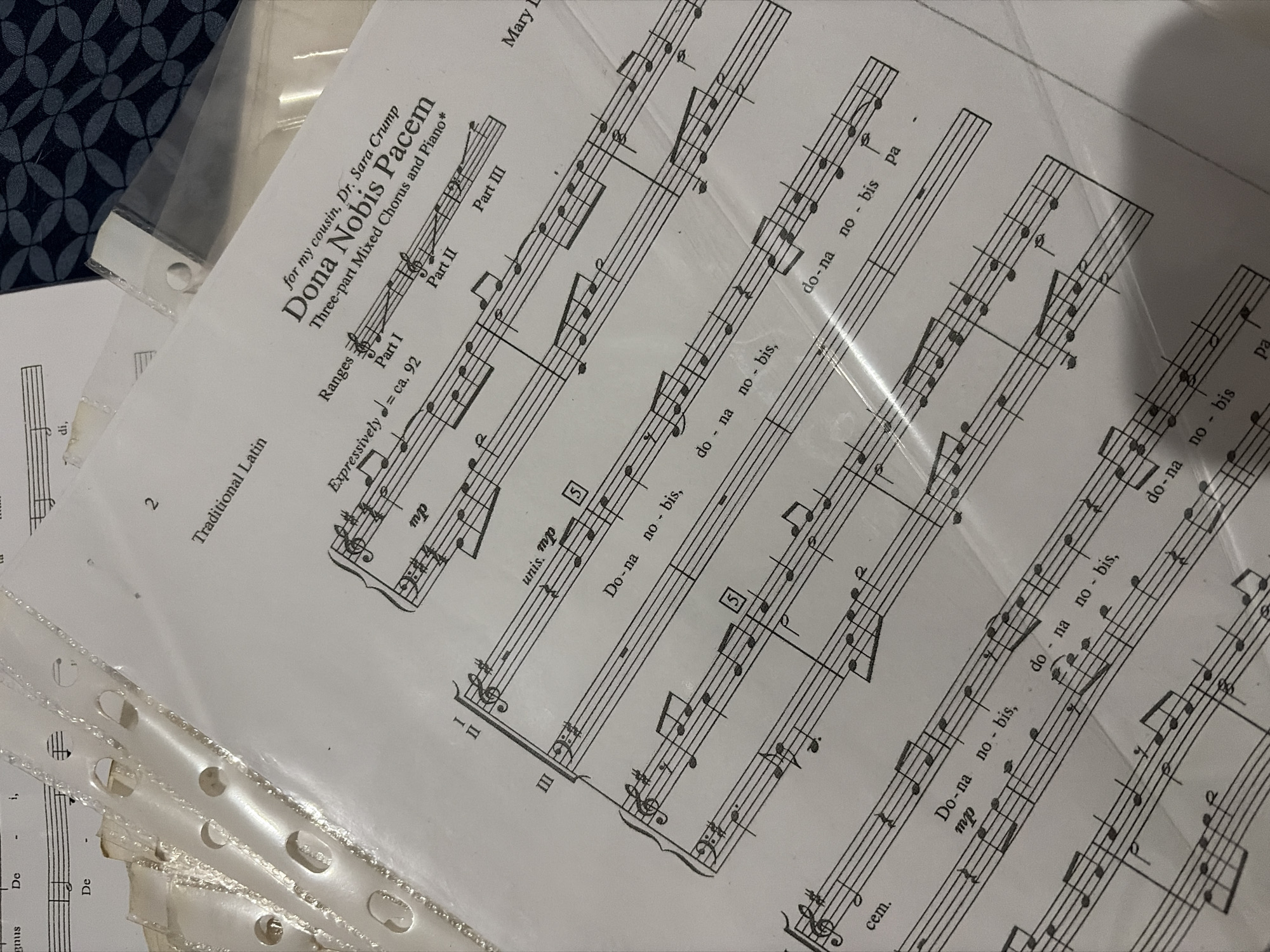 This image, captured in Ciudad Guayana, Venezuela, presents a close-up, angled view of several sheets of musical notation paper, predominantly housed within transparent, perforated plastic sheet protectors. The most prominent sheet, positioned diagonally from the lower left to the upper right, displays the title "Dona Nobis Pacem" for "Three-part Mixed Chorus and Piano," dedicated "for my cousin, Dr. Sara Crump." Text on this sheet includes "Traditional Latin," a page number "2," tempo marking "Expressively d = ca. 92," dynamic markings "mp" and "unis. mp," and sections labeled "Part I," "Part II," and "Part III." Visible musical staves feature treble and bass clefs, notes, rests, and Latin lyrics such as "Do-na no-bis," and "pa-cem." Beneath and partially visible are additional stacked music sheets and plastic sleeves, their punched binding holes along the left edge evident. The background consists of a dark blue or black fabric with a repeating circular or petal-like geometric pattern. No individuals are present, and the scene is static.