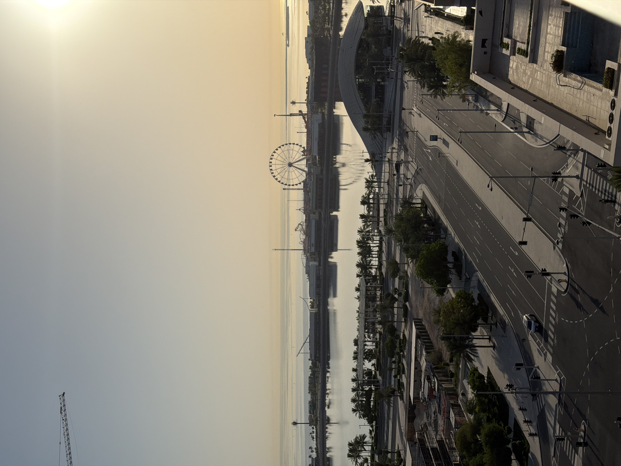 The image shows a wide, elevated view of a waterfront development in an urban setting. The sky is clear with the sun low in the sky, casting long shadows and creating a warm, golden light. In the distance, a large Ferris wheel is visible, along with other amusement park-like structures and buildings bordering a body of water. The water itself is calm and reflects the sky and structures.
In the foreground and middle ground, a modern city street curves through the scene. Palm trees line the sidewalks and some areas are under construction, indicated by scaffolding and construction materials. A white car is stopped at an intersection controlled by traffic lights. Several streetlights are positioned along the road. On the right side of the image, a building with a rooftop pool and some landscaping is visible. The overall impression is one of a developing coastal city, possibly a tourist destination, captured during the early morning or late afternoon.