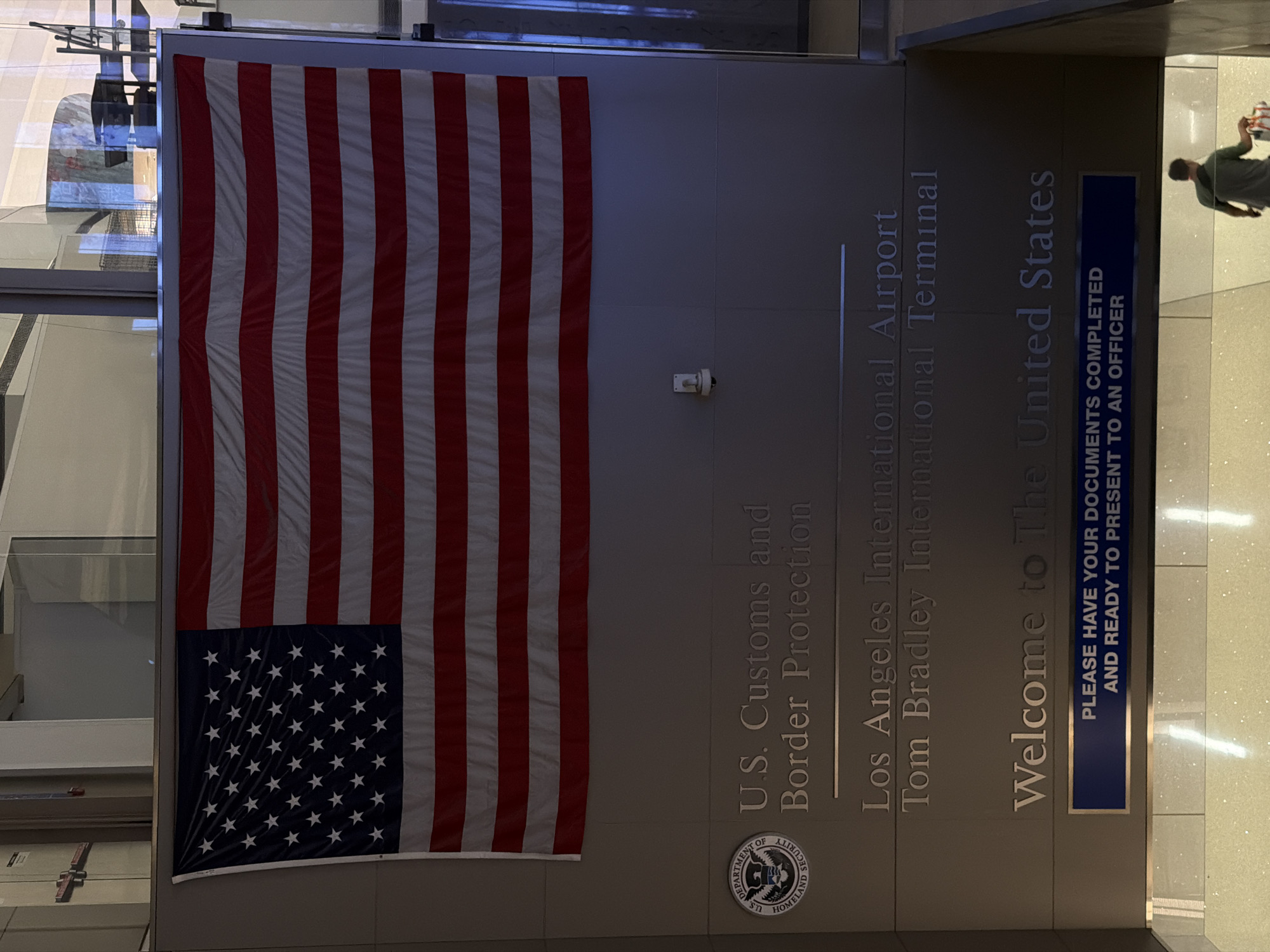 The image shows an interior view of the Tom Bradley International Terminal at Los Angeles International Airport. The primary subject is a large American flag hanging next to a wall bearing the inscription "U.S. Customs and Border Protection," "Los Angeles International Airport Tom Bradley International Terminal," and "Welcome to The United States." A sign in a blue frame reads "PLEASE HAVE YOUR DOCUMENTS COMPLETED AND READY TO PRESENT TO AN OFFICER." A person is walking out of frame, towards the right side.