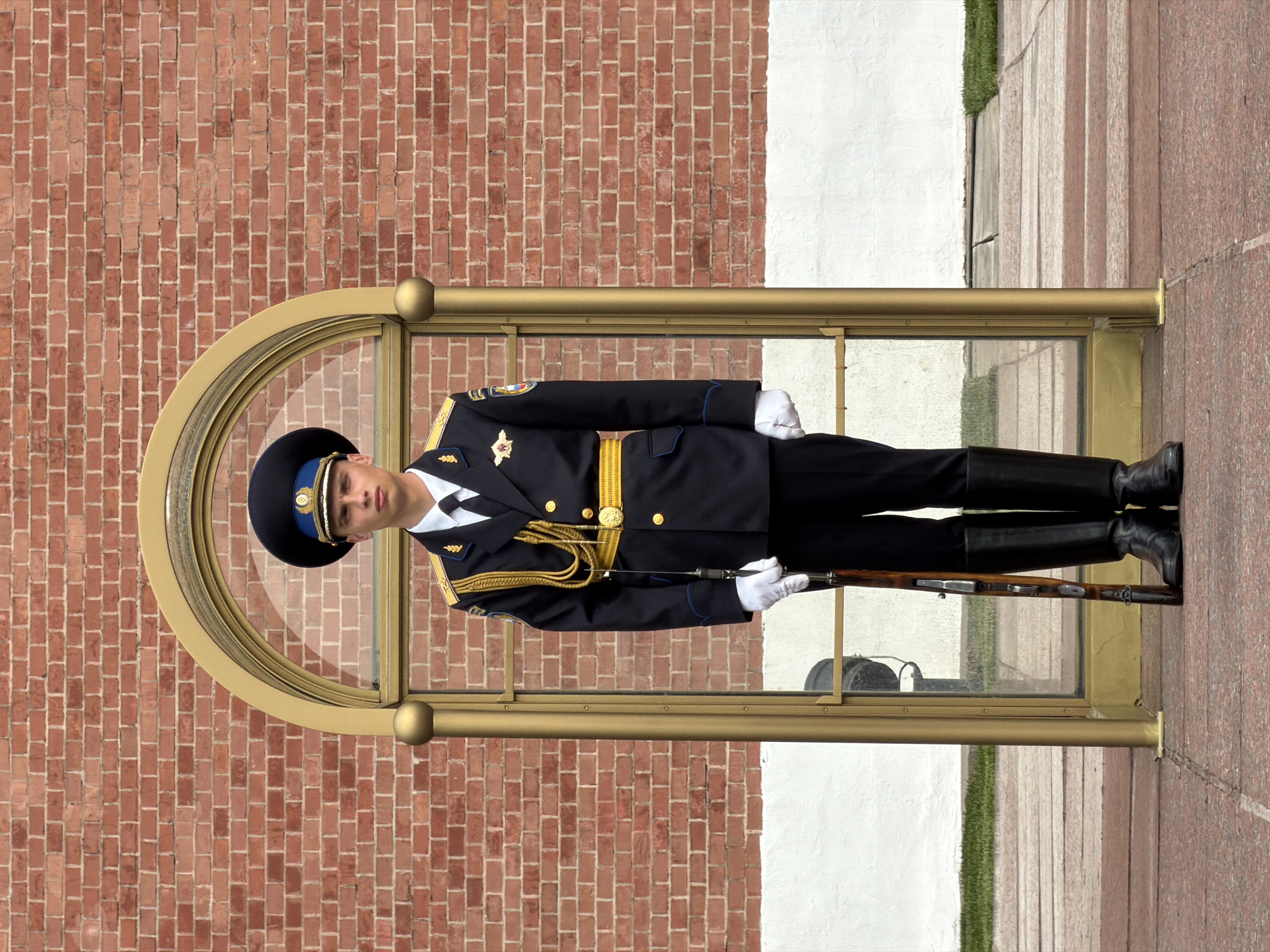 A uniformed sentry stands guard in front of a brick building in Moscow, Russia. The sentry wears a dark blue military uniform, white gloves, and black boots and hat. He holds a rifle. He is standing in a glass enclosure. The brick building is red. There is a granite path to the right of the glass enclosure. There is a white wall and some grass behind the glass enclosure.