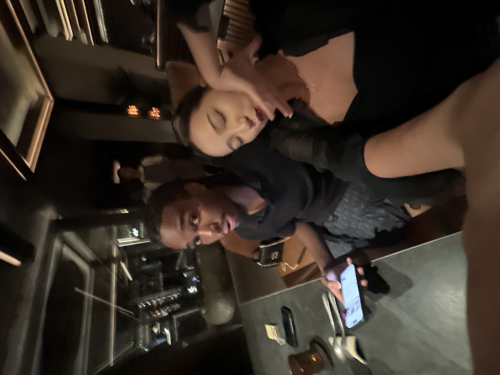The image captures a nighttime scene indoors. A man is holding a smartphone with the screen illuminated. He appears to be taking a picture or recording a video of a woman whose face is being touched by one or two other people. A person is either holding her or has their hands on her neck/shoulders. Cutlery and other objects are present on the table. The location is North Kuta, Indonesia.
