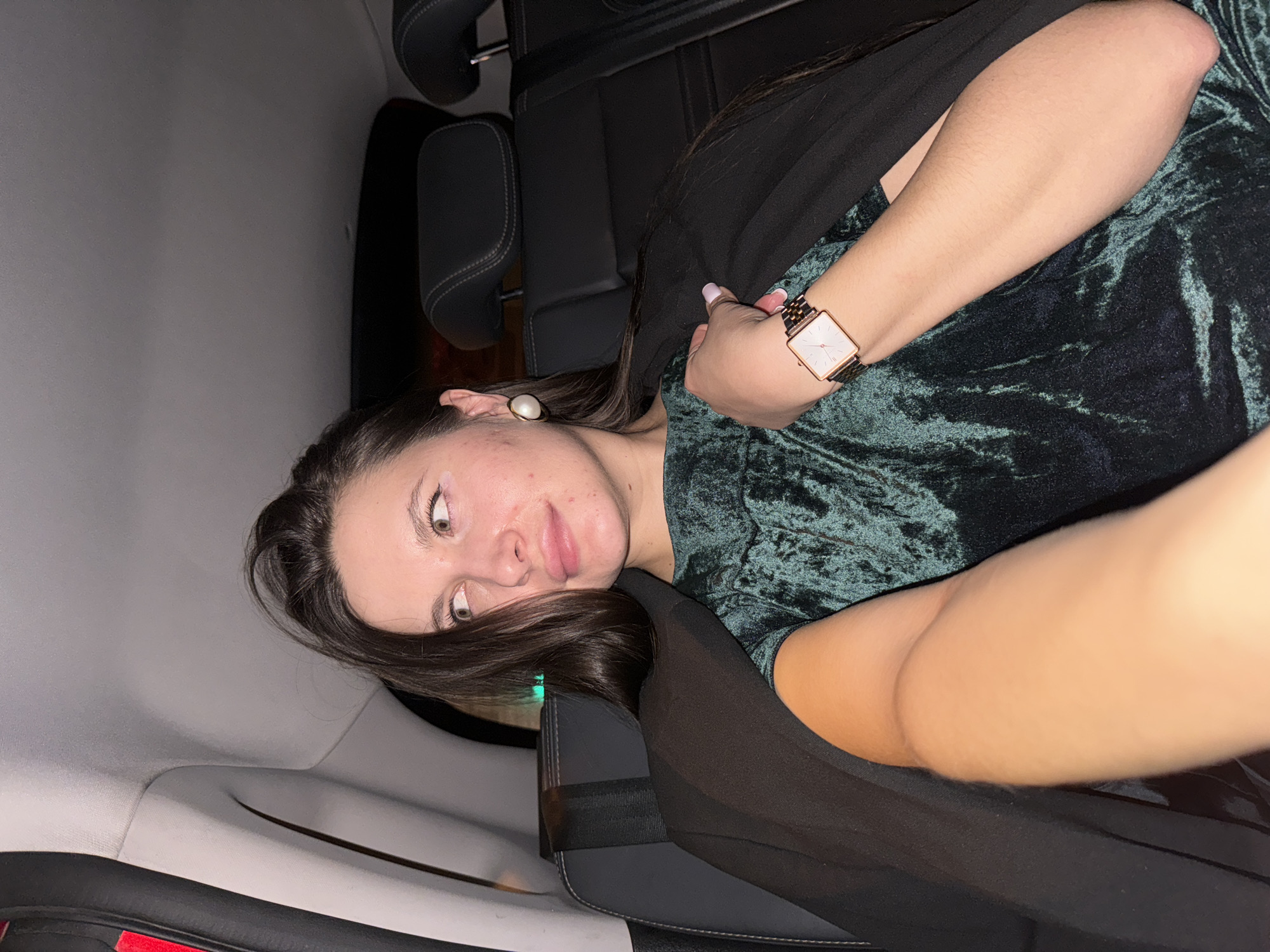 A woman is reclining in what appears to be the back seat of a car. She is wearing a dark green crushed velvet top, a black shawl or jacket, and a rectangular-faced watch with a rose gold finish on her wrist. Her hair is dark and falling across the headrest. She has a pearl earring in her left ear. The car's interior includes black leather seats and gray paneling. The location is unspecified and unknown.