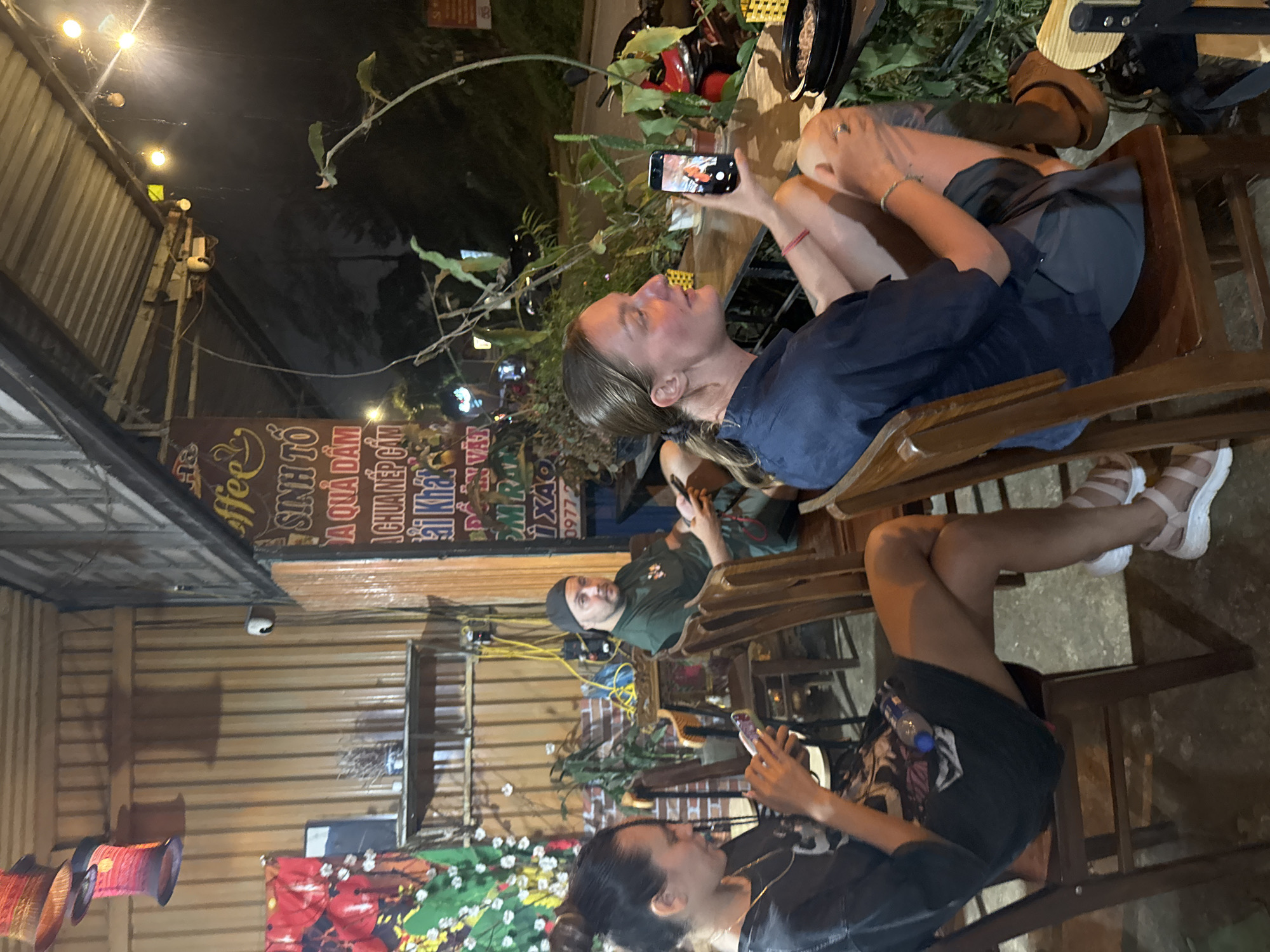 This image captures a night scene at an open-air cafe or restaurant in Mù Cang Chải, Vietnam. Several individuals are seated at wooden tables and chairs, illuminated by string lights and other artificial light sources.
In the foreground, a person with light brown hair, wearing a dark blue shirt and shorts, is seated, holding a smartphone and seemingly taking a picture or video of something on the table or across from them. Their legs are crossed. Across from them, another person with a beard and a dark cap, wearing a green t-shirt, is seated, looking towards the viewer or slightly past them, also holding a phone. Further in the foreground on the right, a third person with dark hair, dressed in a dark t-shirt and shorts, is also engrossed in their smartphone.
The setting features wooden slatted walls, a corrugated metal roof structure, and various potted plants around the seating area, creating a relaxed ambiance. Decorative red lanterns hang on the left side of the frame. Overhead, string lights cast a warm glow. A security camera is visible on one of the wooden pillars. The general mood appears casual and social, with people engaged with their devices or companions.
Visible text on a large sign in the background includes "Coffee" in English, and Vietnamese words such as "SINH TỐ" (smoothie), "A QUẢ ĐẬM" (a type of fruit drink or deep flavor), "CHỮA ĐẮNG" (perhaps relating to a specific coffee or bitter taste), "KHÁI", and a phone number starting with "0977". The darkness surrounding the establishment confirms it is nighttime.
