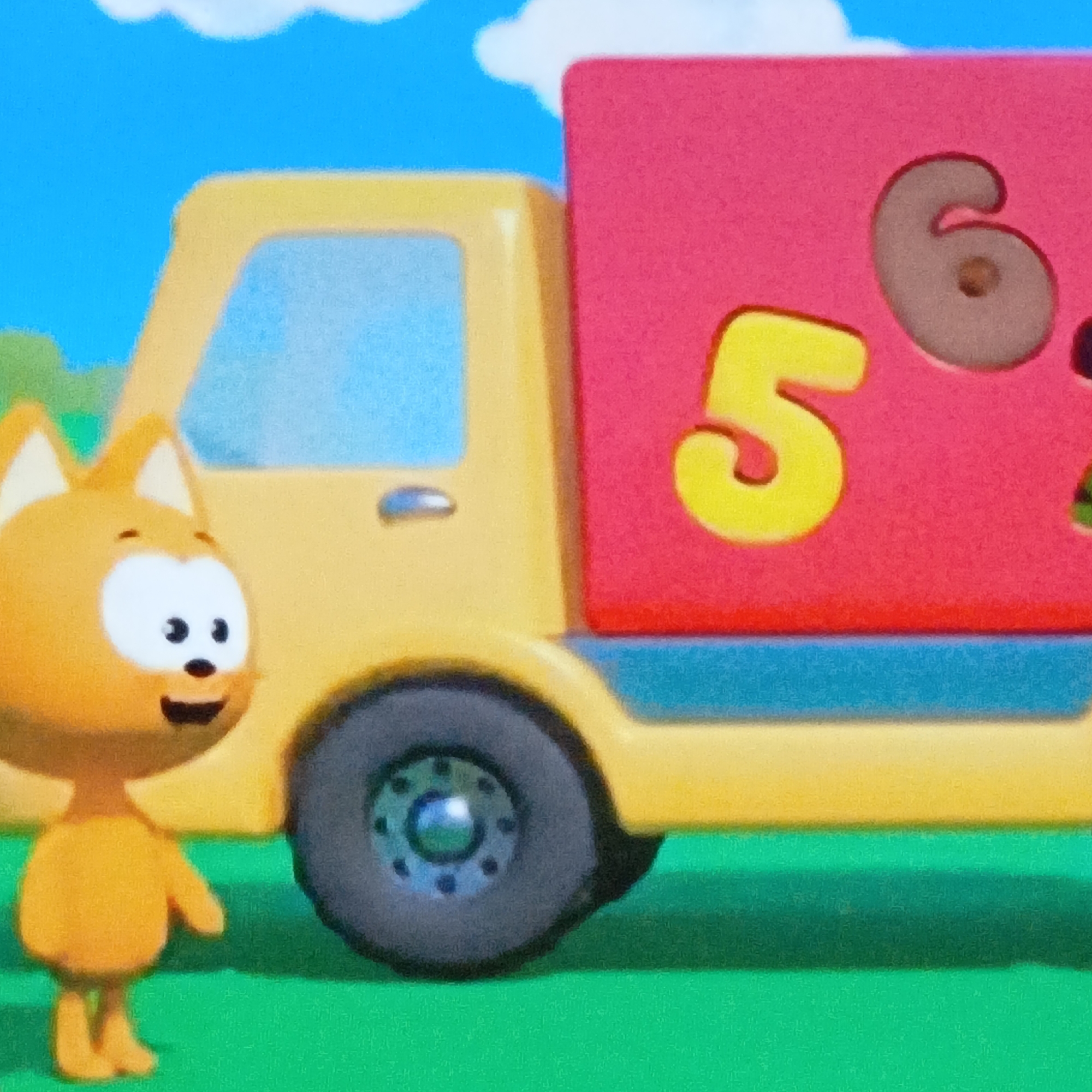 An eye-level, bright, and colorful image depicts a playful, cartoon-like scene. The primary subject is an orange, feline-like character, possibly a cat or fox, standing on the left side of the frame. This character has large white eyes with black pupils, a small black nose, a wide, smiling black mouth, and pointed ears. It wears no clothing and has stubby arms and legs.
To the right of the character is a yellow toy truck with light blue windows and a red cargo bed. A small blue handle is visible on the truck's cab door. The truck has a dark gray wheel with a blue-hued hubcap on the front. On the red cargo bed, there's a red block or box with cut-out shapes for numbers. The numbers "5" (in yellow) and "6" (in a brownish-gray) are prominently displayed.
The setting appears to be a bright, clear day, with a vibrant green ground suggesting grass and a light blue sky dotted with fluffy white clouds in the background. The overall impression is that of a cheerful, animated world, possibly from a children's show or educational toy context, given the numbers on the truck. The scene is brightly lit, with no shadows suggesting a particular time of day beyond daylight. No specific location cues are present beyond the generic outdoor elements.