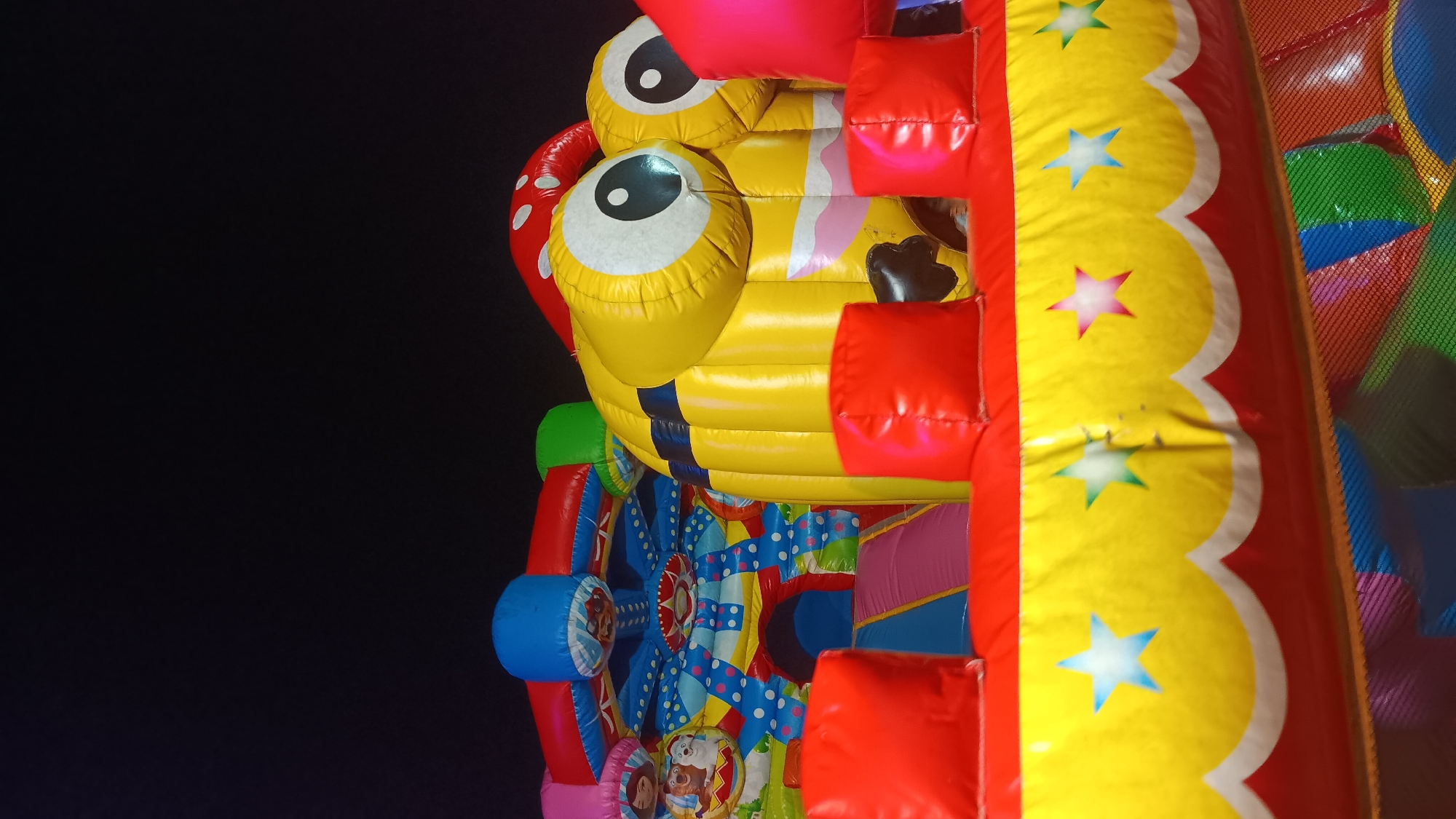 An inflatable playground is set up at night in Getas, Indonesia. The playground features a large yellow Minion character with two eyes, red mushroom-shaped decorations, and a Ferris wheel-like structure with colorful cartoon characters. The foreground shows a red and yellow inflatable barrier with stars and wavy patterns. The background is dark, indicating it is nighttime. No people are visible in the image.