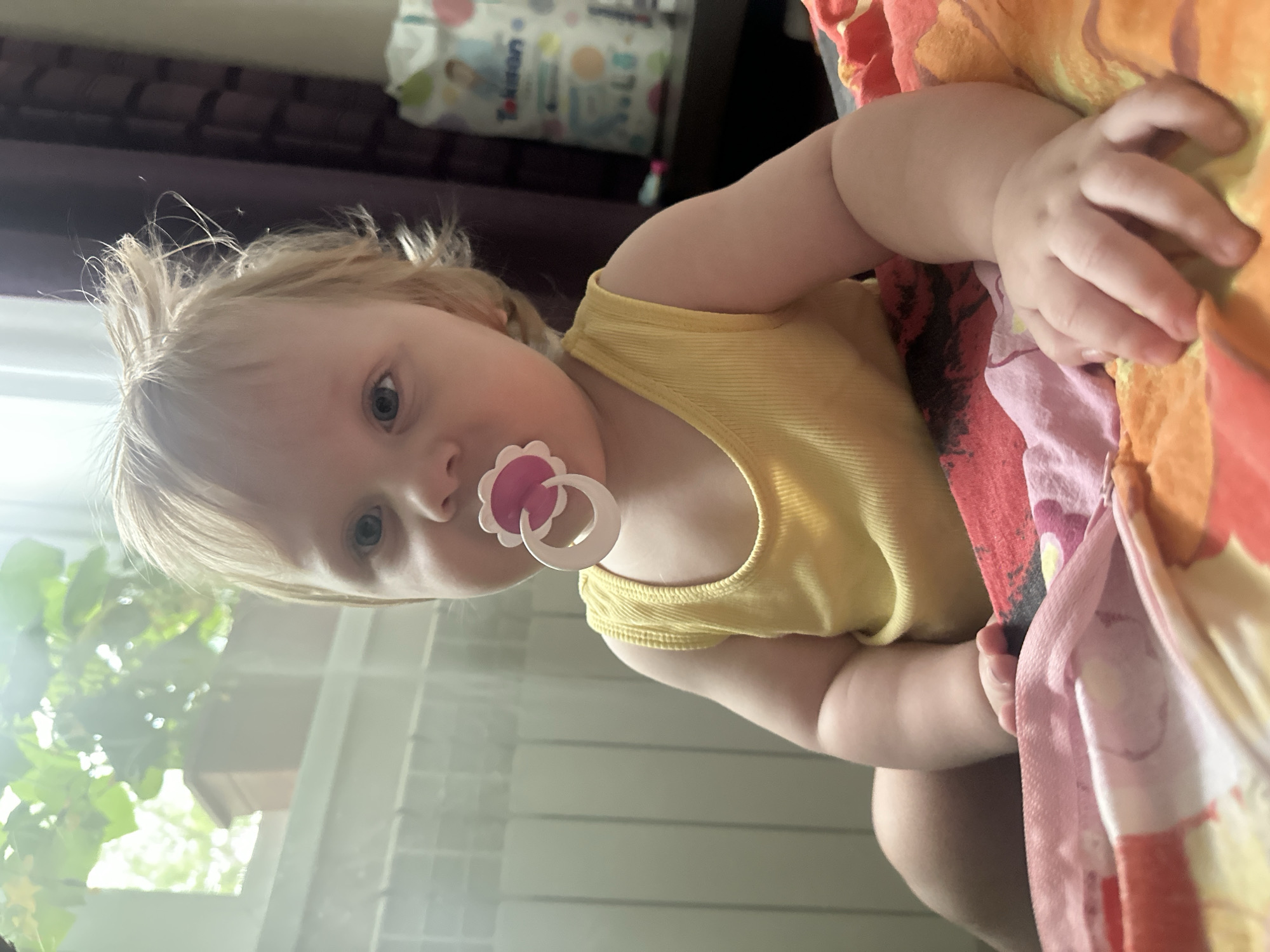 The image shows a young child with blond hair and blue eyes, wearing a yellow tank top and a pink and white pacifier. The child is sitting on a colorful bedspread, with a radiator and a window with plants in the background. A bag of diapers is visible on a shelf to the right. The scene appears to be indoors, likely in a residential setting. The image does not provide any specific information to determine its location in the city of Sayanogorsk, Russia.
