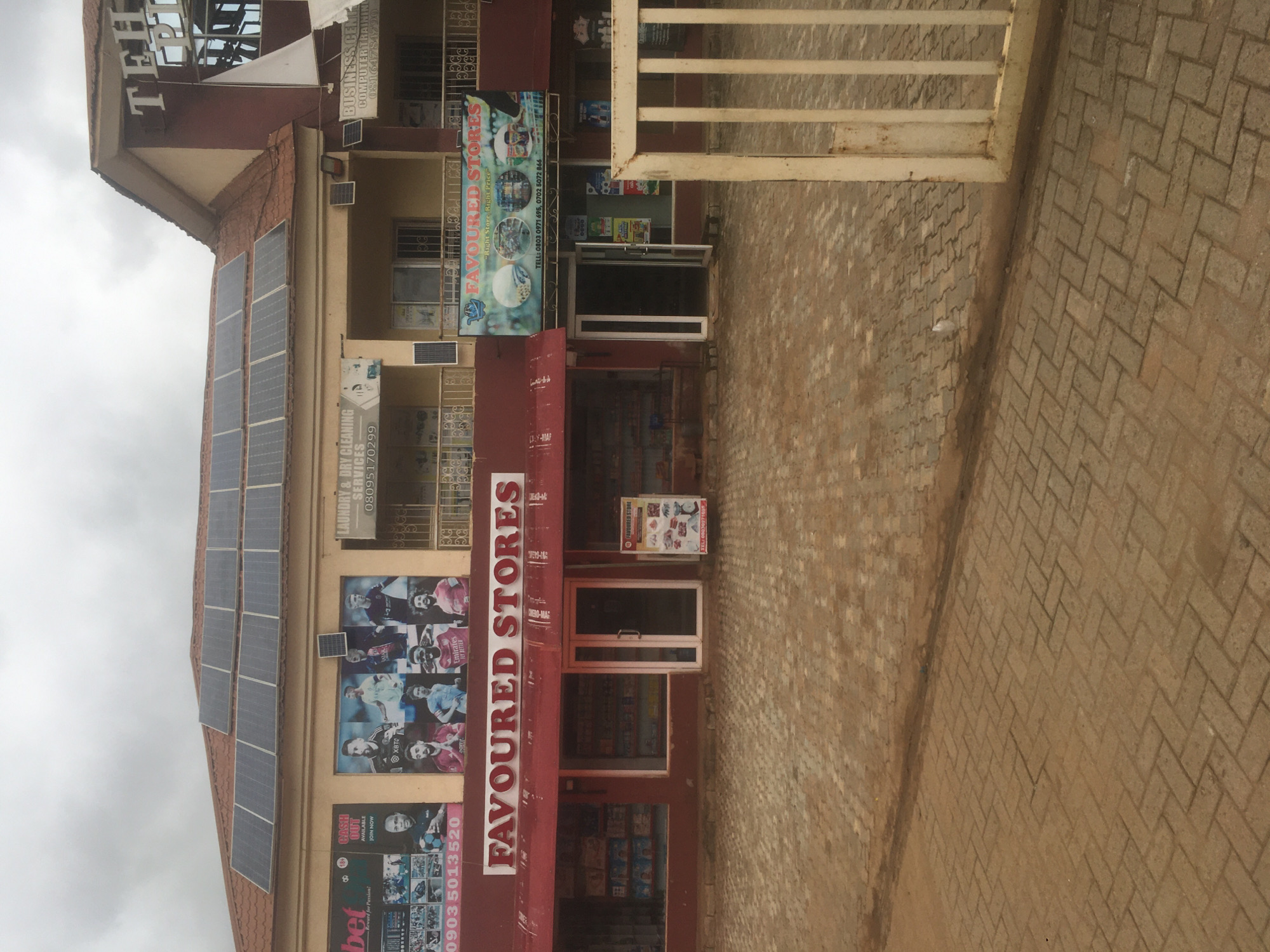 This eye-level, medium shot captures the exterior of a multi-story commercial building in Dutsen Alhaji, Nigeria, under an overcast sky. The building, featuring a terracotta-colored roof and light brown walls, houses several businesses on its ground floor. Numerous solar panels are installed across the roof.
The most prominent business is "FAVOURED STORES," indicated by large red signage. One sign includes the slogan "Right Store, Right Price" and phone numbers: "0803 0971 695" and "0702 5072 866." Adjacent to it is a betting shop with a large banner displaying sports figures and the text "bet Reward for Passion! 18 CASH OUT AVAILABLE JOIN NOW 09035013520." Another smaller sign shows "Emirates FLY BETTER." Other businesses visible include "LAUNDRY & DRY CLEANING SERVICES" with phone number "08095170299," and a "BUSINESS CENTRE COMPUTER TRAINING" with phone number "08164784790."
The ground in front of the building is paved with light brown interlocking bricks. A rustic, white metal gate or barrier partially obstructs the right side of the view. No specific human activity is depicted; the scene focuses on the commercial facades and infrastructure. The overall impression is that of a bustling commercial area.
