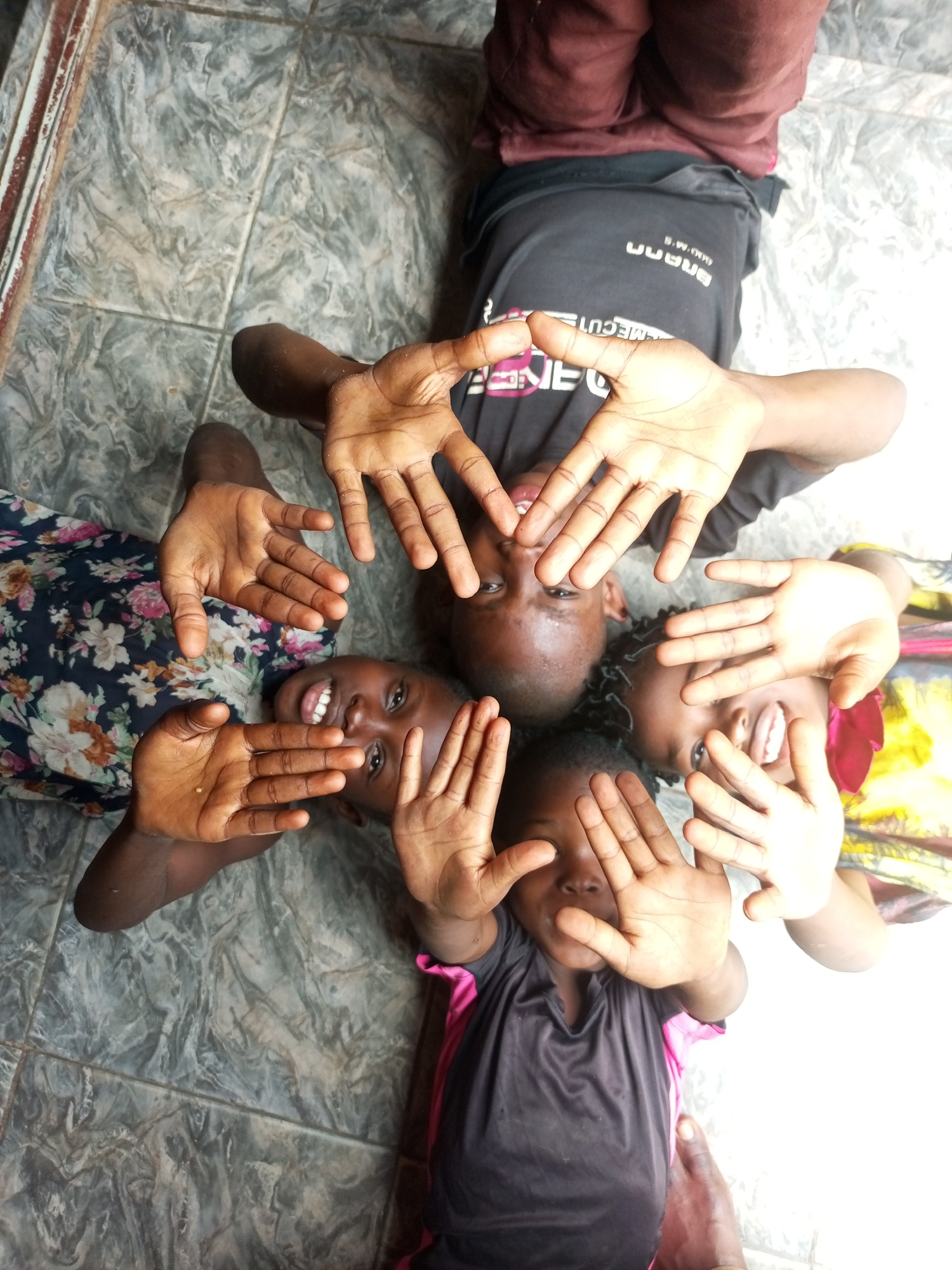 In Gombe, Nigeria, a group of five children lie on a tiled floor, arranged in a circular pattern with their heads towards the center and feet facing outwards. Their hands are raised, partially obscuring their faces. The children are wearing casual clothing, including t-shirts and a floral-patterned dress. The lighting suggests this image was taken indoors or in a shaded outdoor area. The overall scene depicts a moment of playful interaction and shared experience among the children.