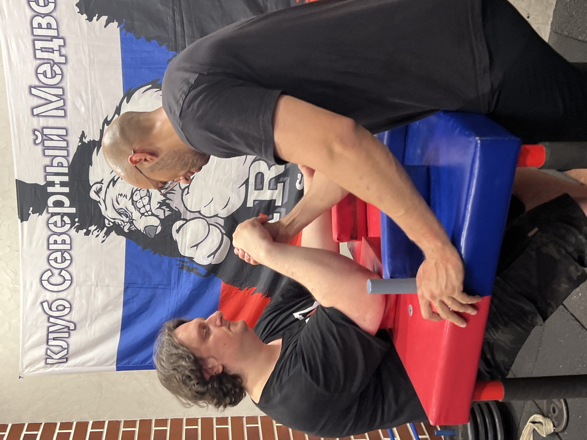 The image depicts two men arm wrestling. They are positioned at a red and blue arm wrestling table. Both men are wearing black t-shirts and appear to be engaged in a competition. The background features a banner with Russian text and a graphic of a bear, suggesting a Russian setting, potentially a club or event related to arm wrestling. The lighting appears to be indoor and artificial. One man has curly brown hair and is looking down at his hands. The other man is bald with glasses and is also focused on the match. The environment seems to be a gym or training facility, with weights visible in the lower left corner. There is no specific indication of the location within Moscow, Russia, beyond the presence of the Russian flag and text.
