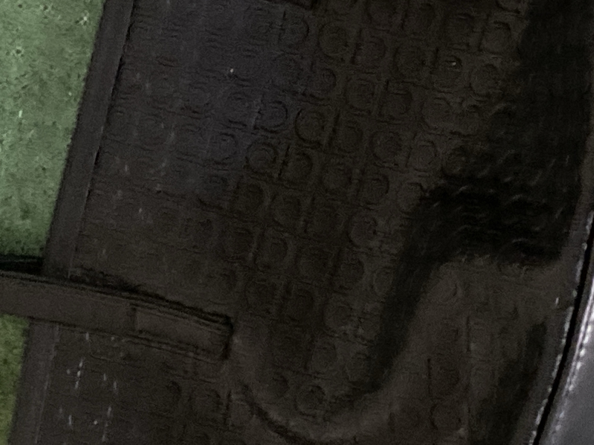A dark, patterned tote bag is pictured in an outdoor setting, with a blurred green surface in the background. The bag appears to be made of patent leather and features a repeating embossed pattern. A dark strap is visible on the upper left side of the bag. The lighting suggests it is daytime, and the image is likely a close-up of the bag. There is no indication of the location in Jauro Yinu, Nigeria.