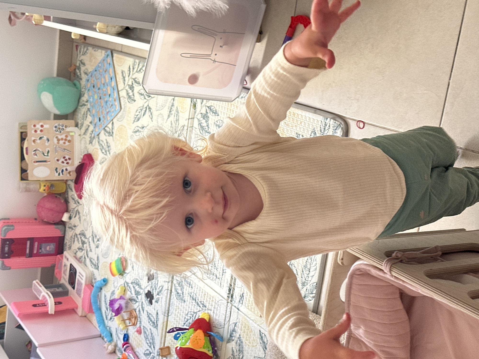 A blond toddler with blue eyes and fair skin, exhibiting a minor red mark on the right cheek, stands on a tiled floor in what appears to be a playroom. The child is wearing a cream-colored, ribbed long-sleeved shirt and green pants. The background reveals a colorful play area with various toys, including a pink toy kitchen, a pink suitcase, a pink ball, a blue stuffed animal, and a number puzzle. A play mat with a leaf pattern is visible on the floor. The environment suggests a domestic setting, possibly a nursery or playroom, located in Petah Tikva, Israel. The child's pose and outstretched hands indicate an interaction with the viewer or an object just out of frame.
