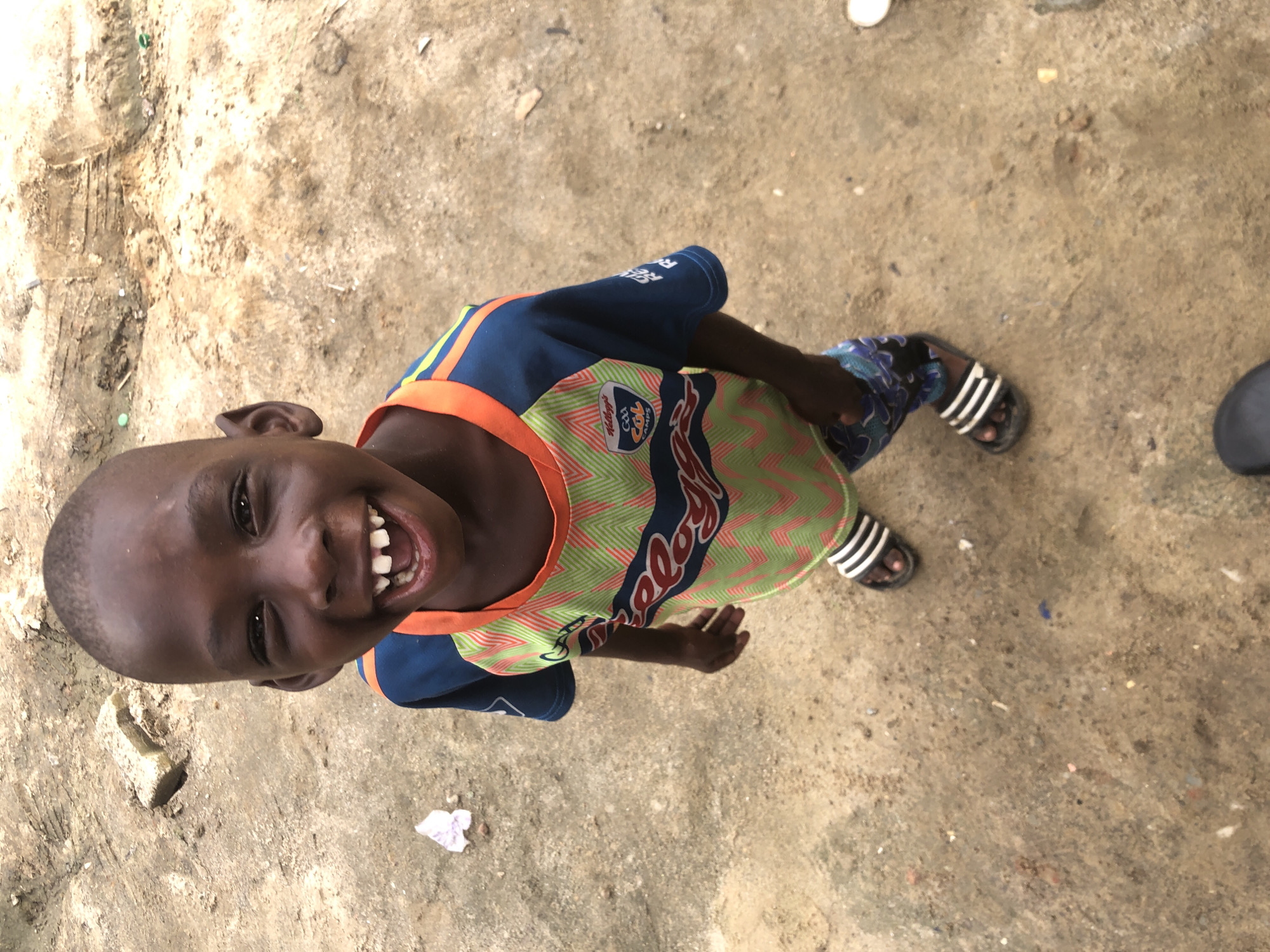 A young Black boy, likely under ten years old, stands on a dirt ground in what appears to be an outdoor urban setting in Ado, Nigeria. The boy is captured from a high angle, looking directly at the camera with a wide, toothy grin. He has dark skin, a shaved head, and his ears are visible. He is wearing a blue and orange t-shirt with a green and lime zigzag pattern across the chest, featuring a logo with "Kellogg's GAA CÚL CAMPS" text and an emblem. He is also wearing blue shorts with a dark pattern and black and white striped slides on his feet. The ground is composed of loose dirt, with scattered small stones, debris, and a few pieces of white and colored paper. In the background, there are mounds of earth and a shallow trench, suggesting a construction or undeveloped area.