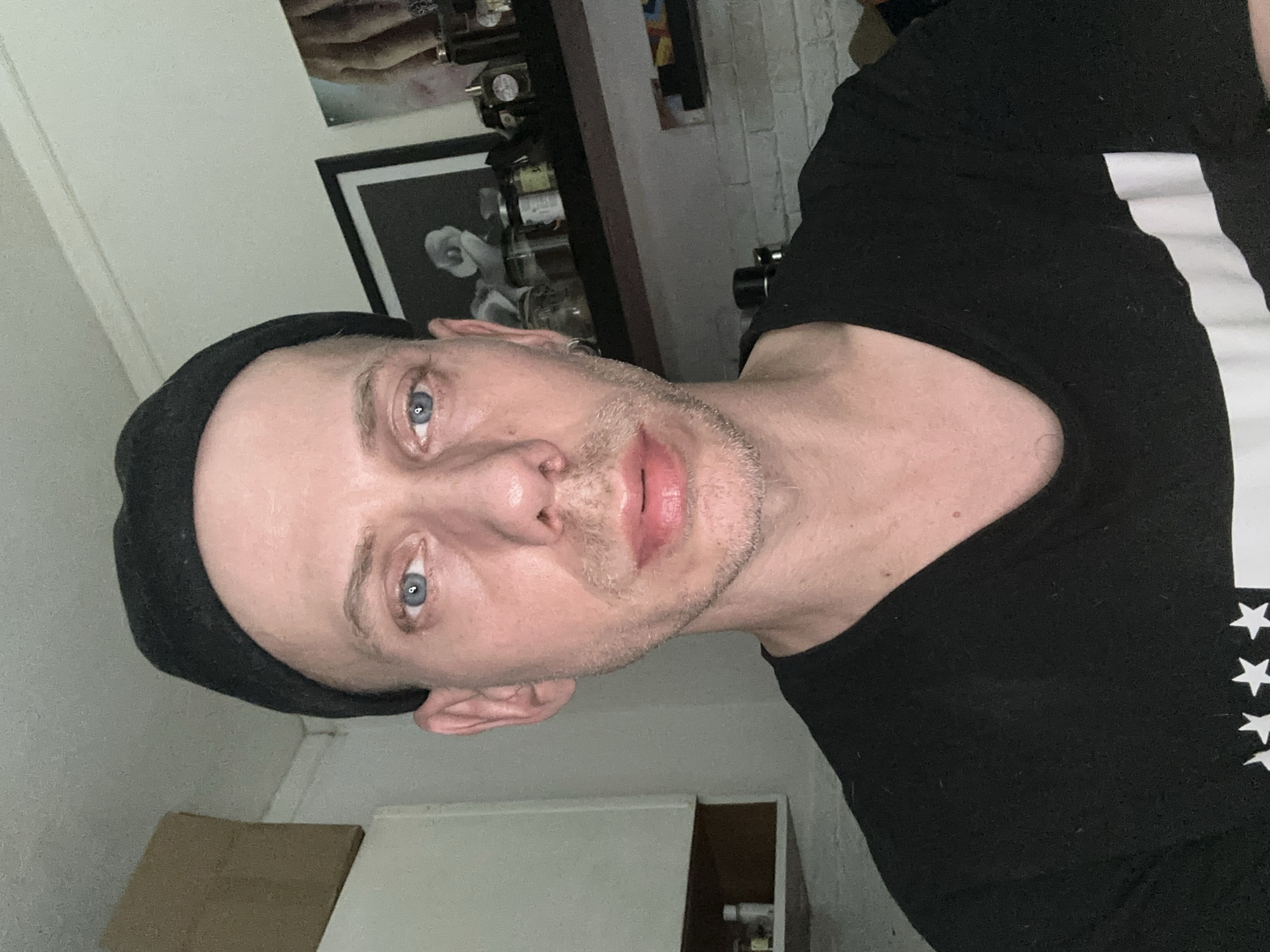 A medium shot shows a fair-skinned person wearing a black beanie and a black t-shirt. The person's eyes are a bright blue and they have light stubble on their chin. The background appears to be an indoor setting with shelves and framed pictures. There is no discernible location in Moscow, Russia.