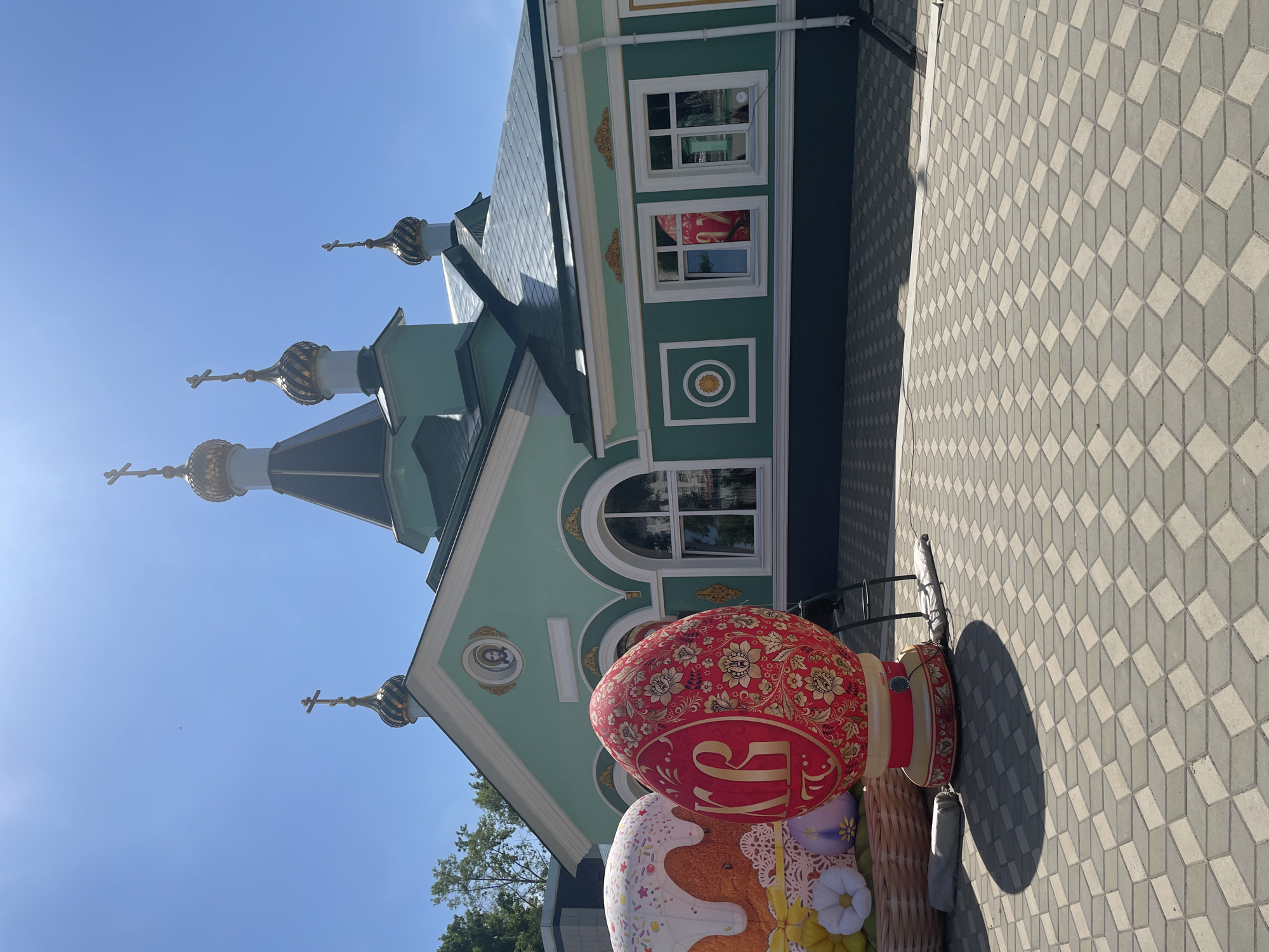 The image shows an eye-level, exterior shot of a building with Russian Orthodox architecture and large Easter decorations, likely taken in Chaikovsky, Russia. The building has a light green facade with white window frames and gold accents. It is topped with three onion domes adorned with gold crosses. To the right of the building is a gray paved area with a cube pattern.
In the foreground, there is an installation of oversized Easter eggs and a traditional Easter bread (kulich) decoration. The large red egg has a gold floral design. The Easter decorations are placed in what appears to be a woven basket.