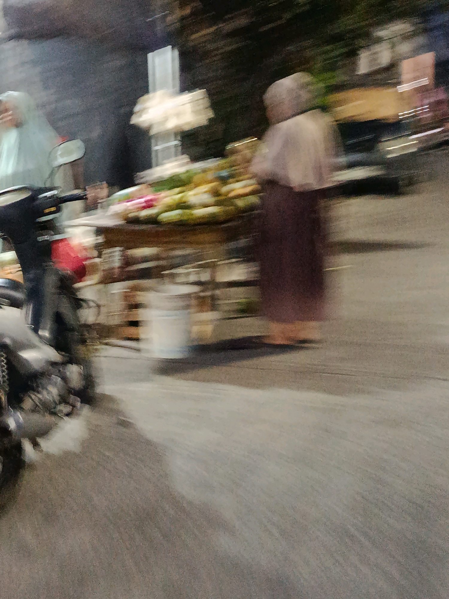 This is a motion-blurred image taken at night in Bekasi, Indonesia, capturing a street scene. In the foreground on the left, a motorcycle is partially visible. A person in a light-colored hijab and dark pants is standing next to the motorcycle. In the middle ground, there is a street vendor's stall with a variety of fruits and vegetables displayed. Behind the stall, a person wearing a darker headscarf and a brown garment stands. The background is obscured by darkness and motion blur, but appears to be an urban setting with some structures. The overall impression is of a bustling street market or roadside activity at night.