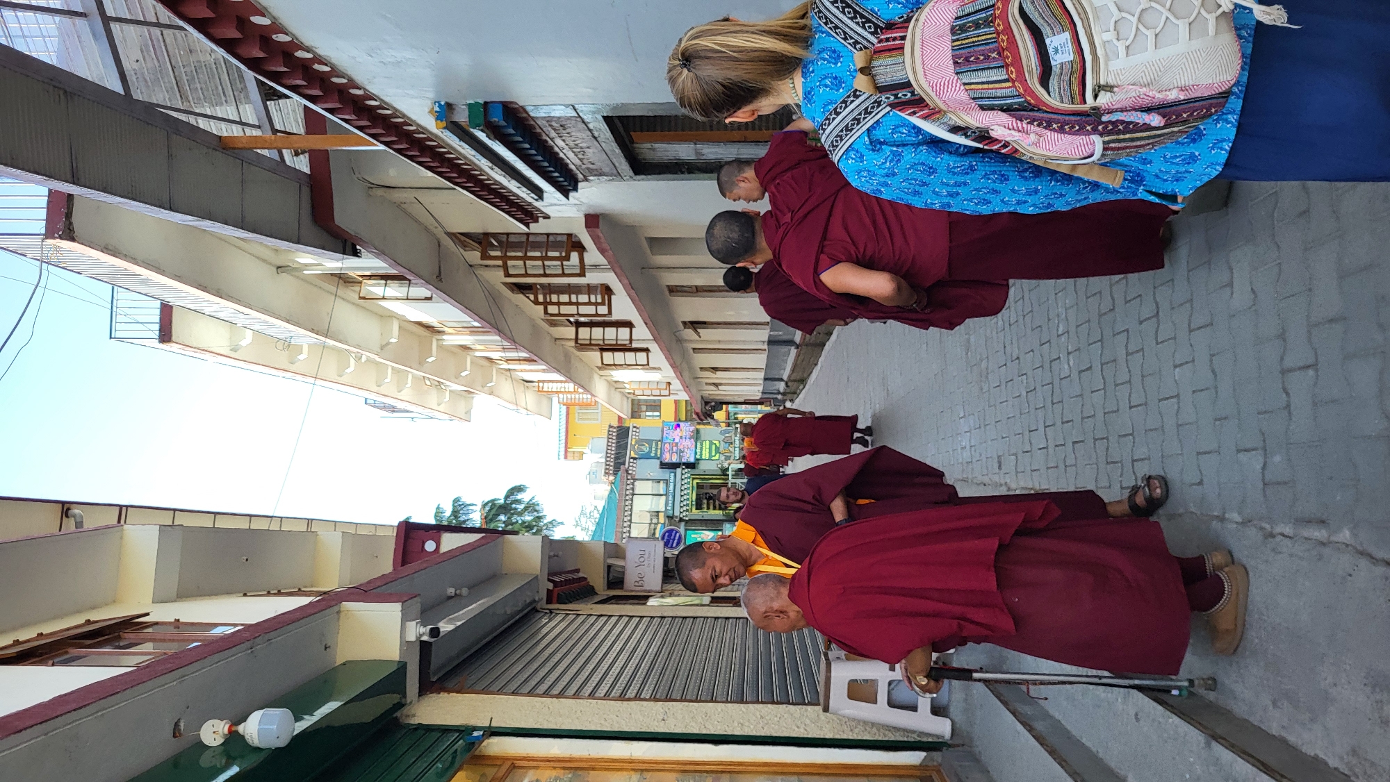 The image depicts a group of Buddhist monks walking along a street in Dharamshala, India. The monks are wearing traditional maroon robes. A woman with blonde hair is standing near the monks, wearing a blue patterned shirt and carrying a striped backpack. The street is lined with buildings, some of which have shops with signs and roll-down metal doors. The buildings are painted in light colors. The street is paved with grey brick. The sky is visible at the end of the street. The camera is oriented to show the street at an upward angle.