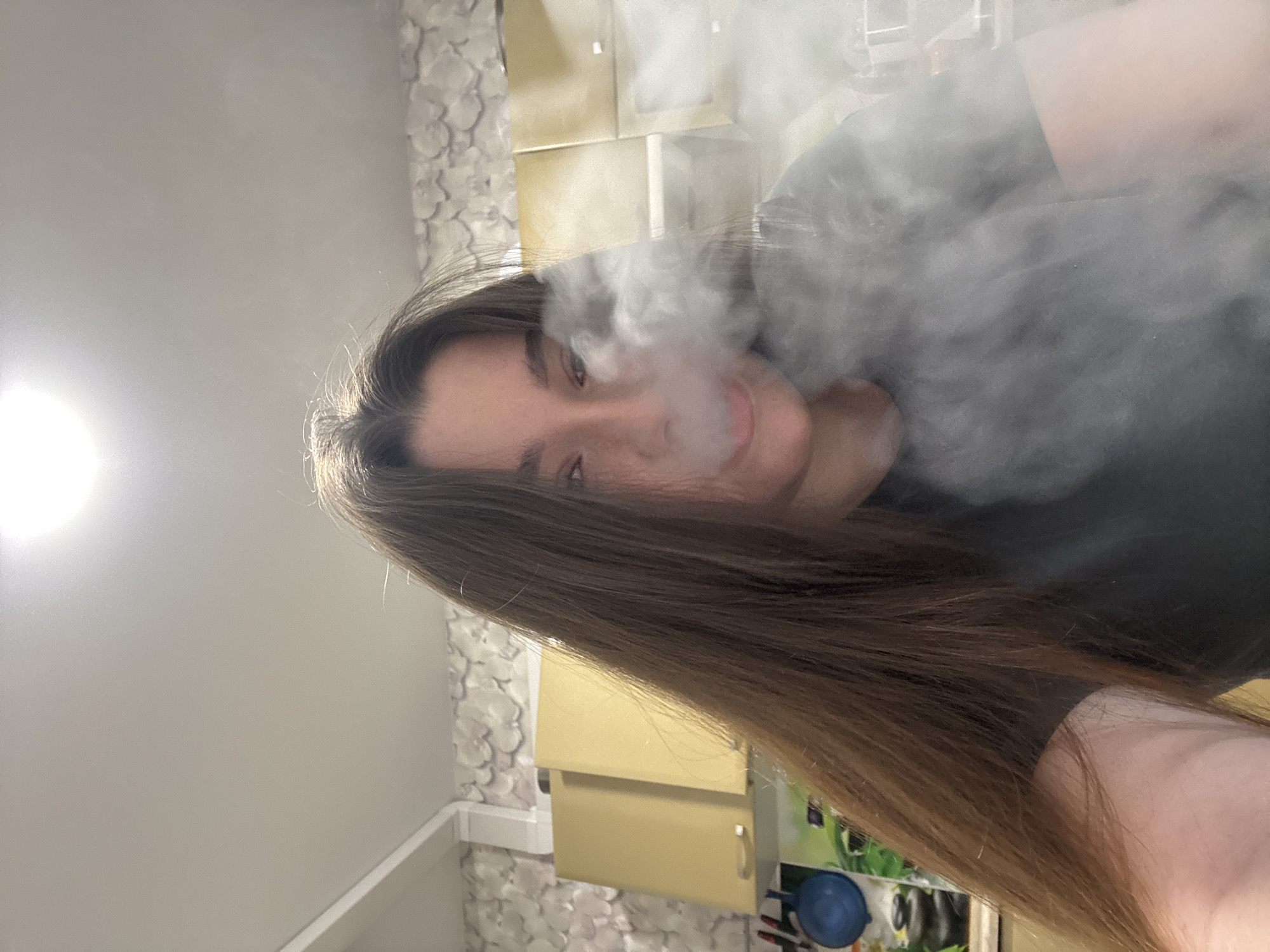 A selfie shot of a woman exhaling a cloud of vapor. She has long, straight, brown hair, and is wearing a dark-colored shirt. The background includes a bathroom setting with white patterned wallpaper and yellow cabinets. The location might be an apartment in Perm, Russia, based on general aesthetic clues and the potential origin of the image.