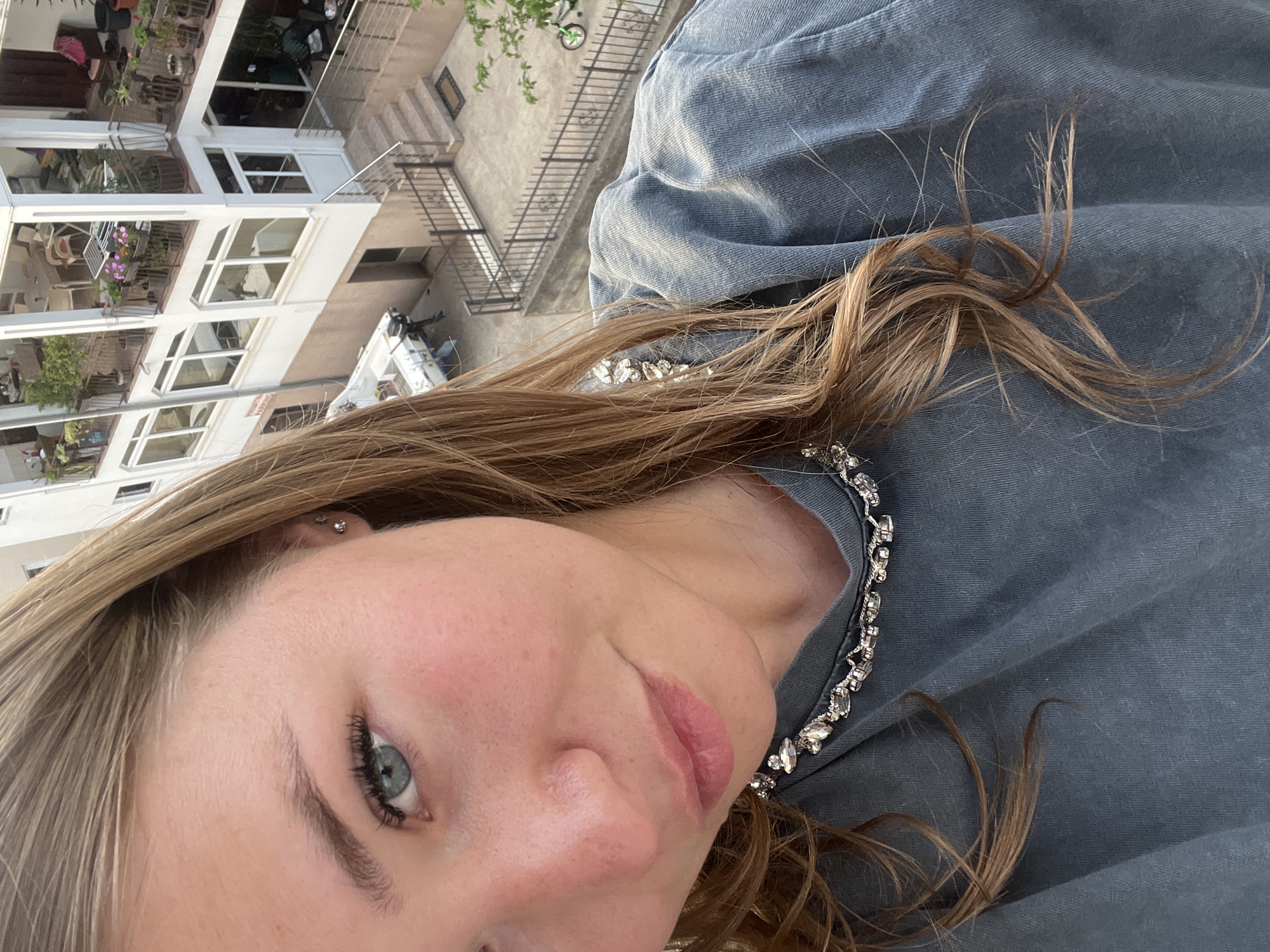 A selfie photograph depicts a fair-skinned woman with light brown hair and blue eyes. She is wearing a blue garment adorned with a bejeweled collar. The background reveals an urban setting in Budva, Montenegro, featuring multi-story buildings with white facades and balconies. A street and sidewalk are visible, and a bicycle leans against a railing.